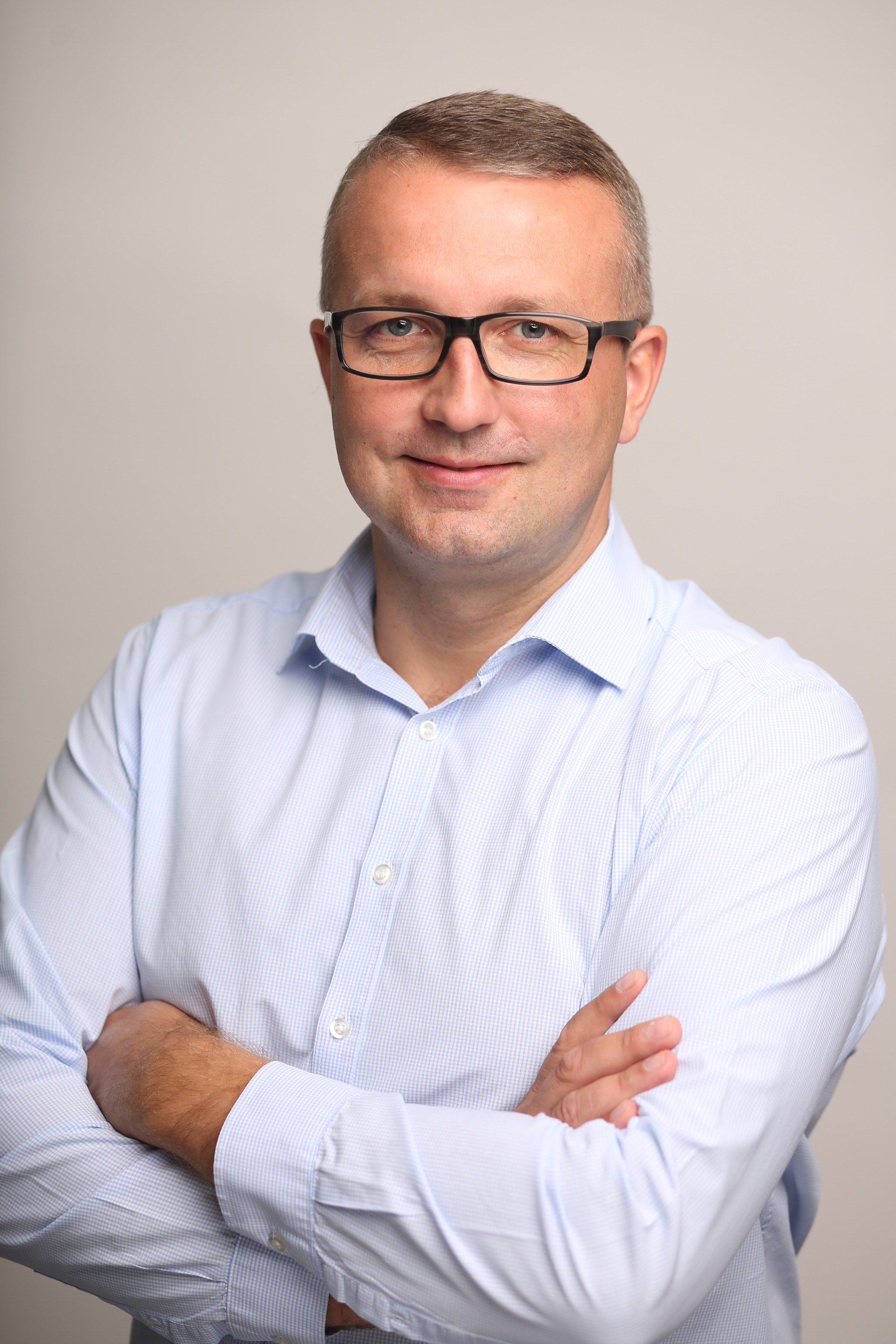 Portret mężczyzny w okularach i jasnej koszuli z założonymi rękami na neutralnym tle.
