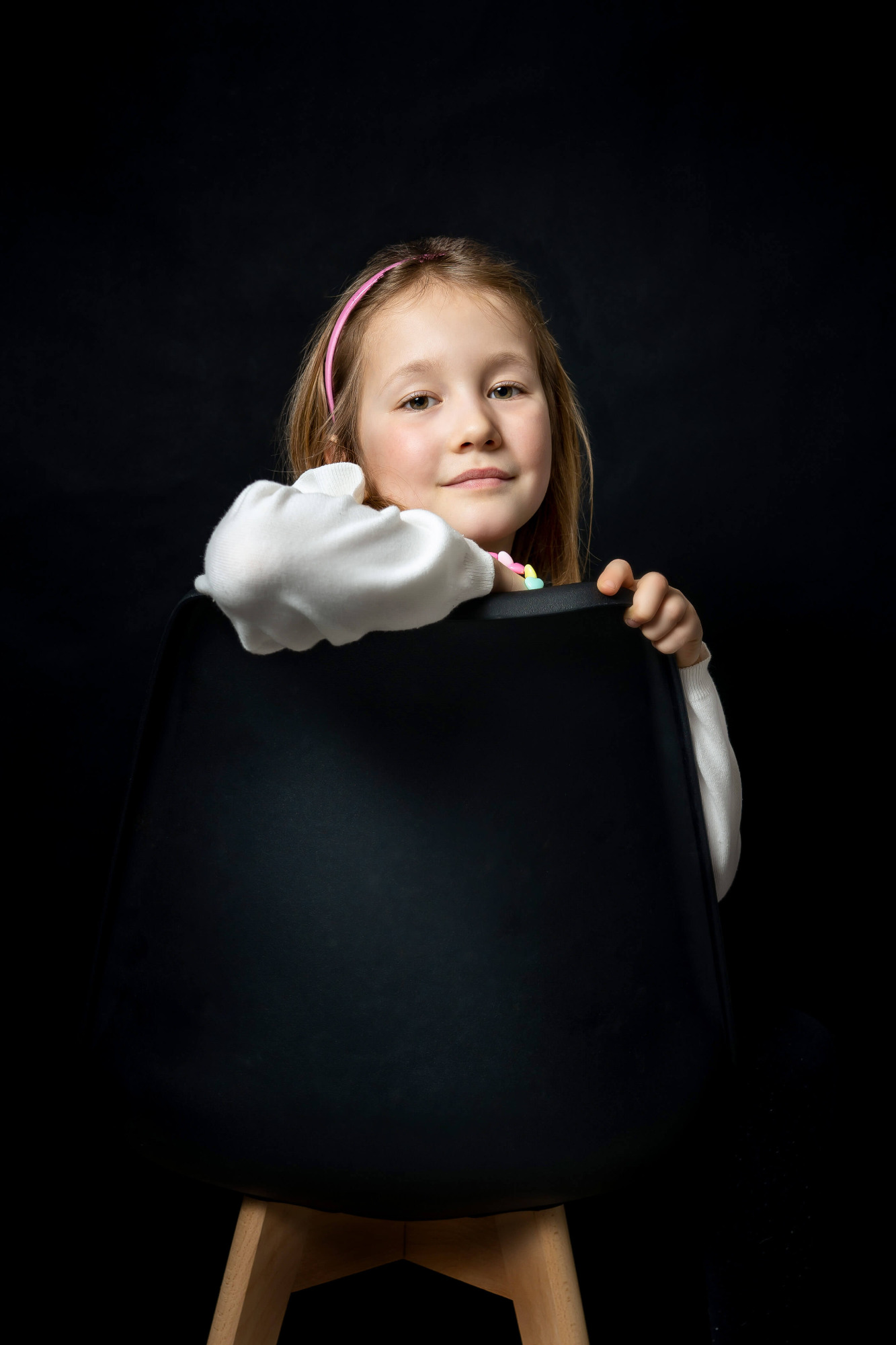 Portret uśmiechniętej dziewczynki z różową opaską, opierającej się o czarne krzesło na ciemnym tle.