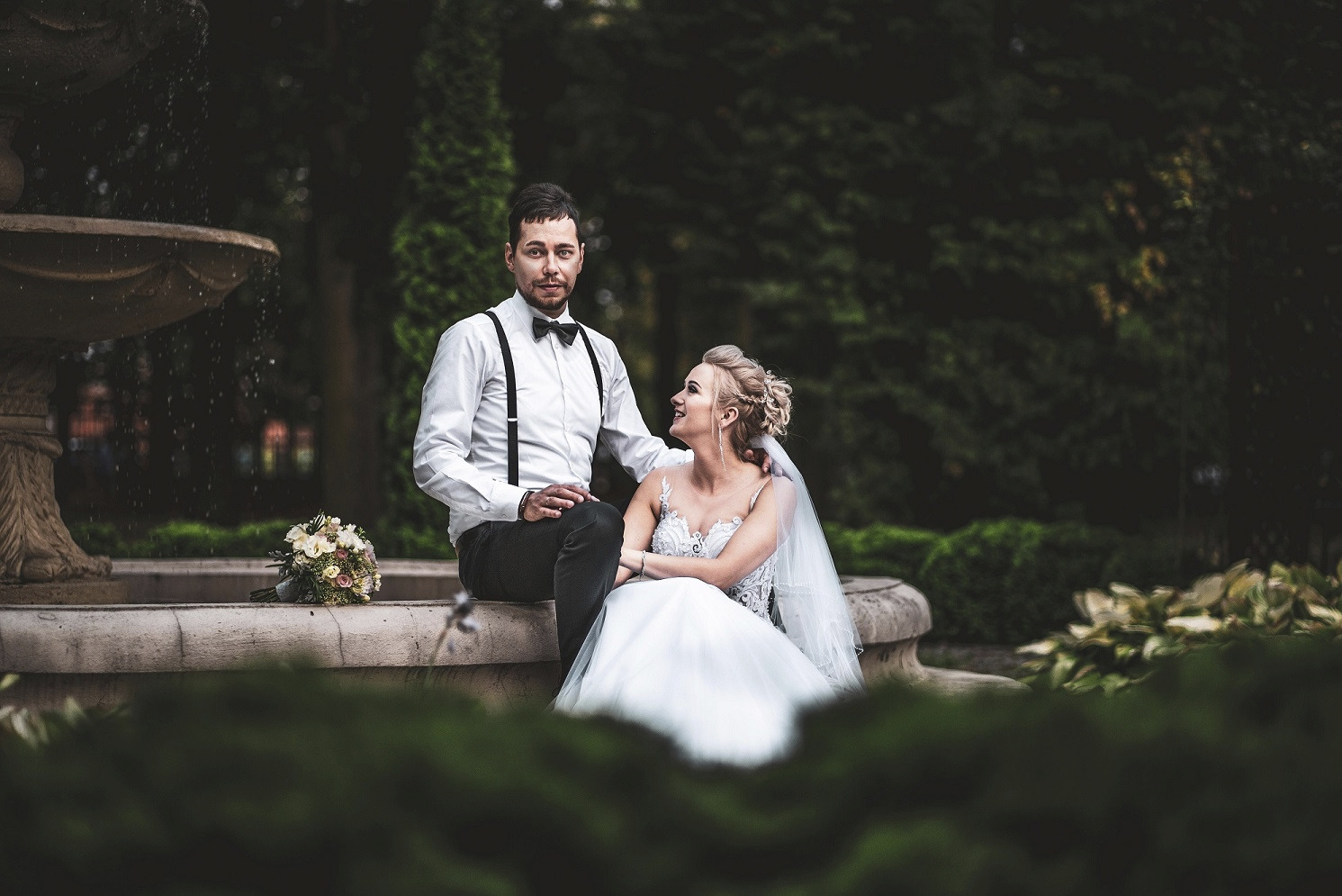 Para młoda siedząca na fontannie: panna młoda w białej sukni z koronkowym gorsetem i welonem, pan młody w białej koszuli z muchą i szelkami, bukiet ślubny obok, ciemne tło z zielenią.