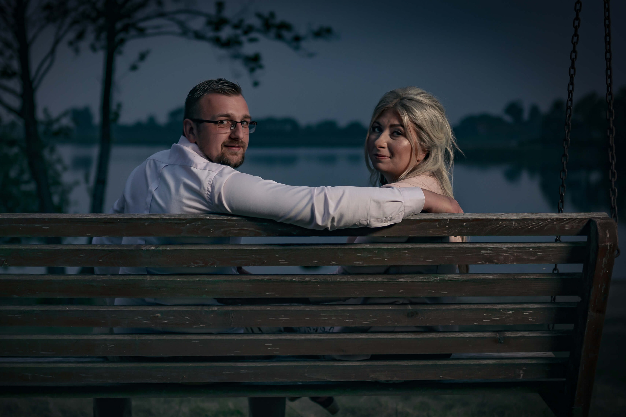 Para siedząca na drewnianej huśtawce nad jeziorem o zmierzchu, mężczyzna w białej koszuli i okularach, kobieta z blond włosami, romantyczna sesja.