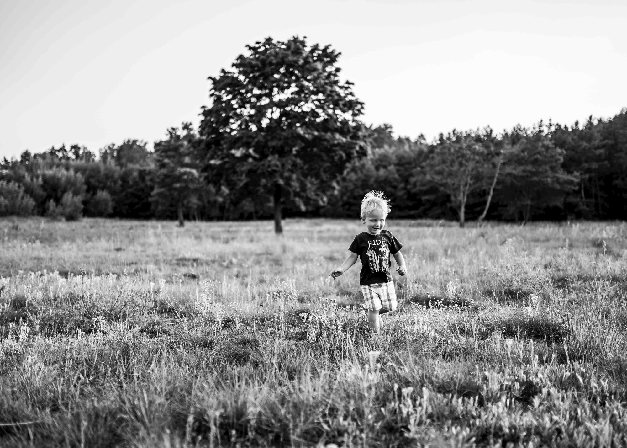 Czarno-białe zdjęcie uśmiechniętego chłopca biegnącego przez łąkę w okolicach Chodzieży, z drzewem w tle.