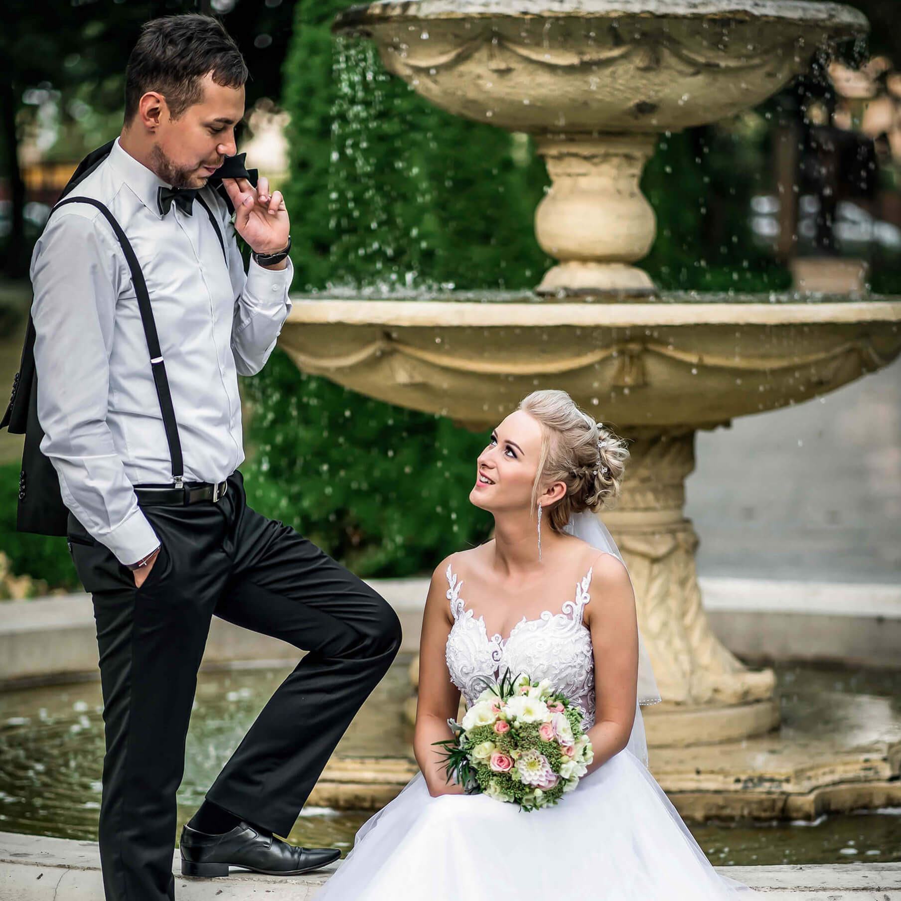 Para młoda w strojach ślubnych pozuje na tle fontanny: panna młoda siedzi z bukietem, pan młody stoi obok z marynarką na ramieniu.