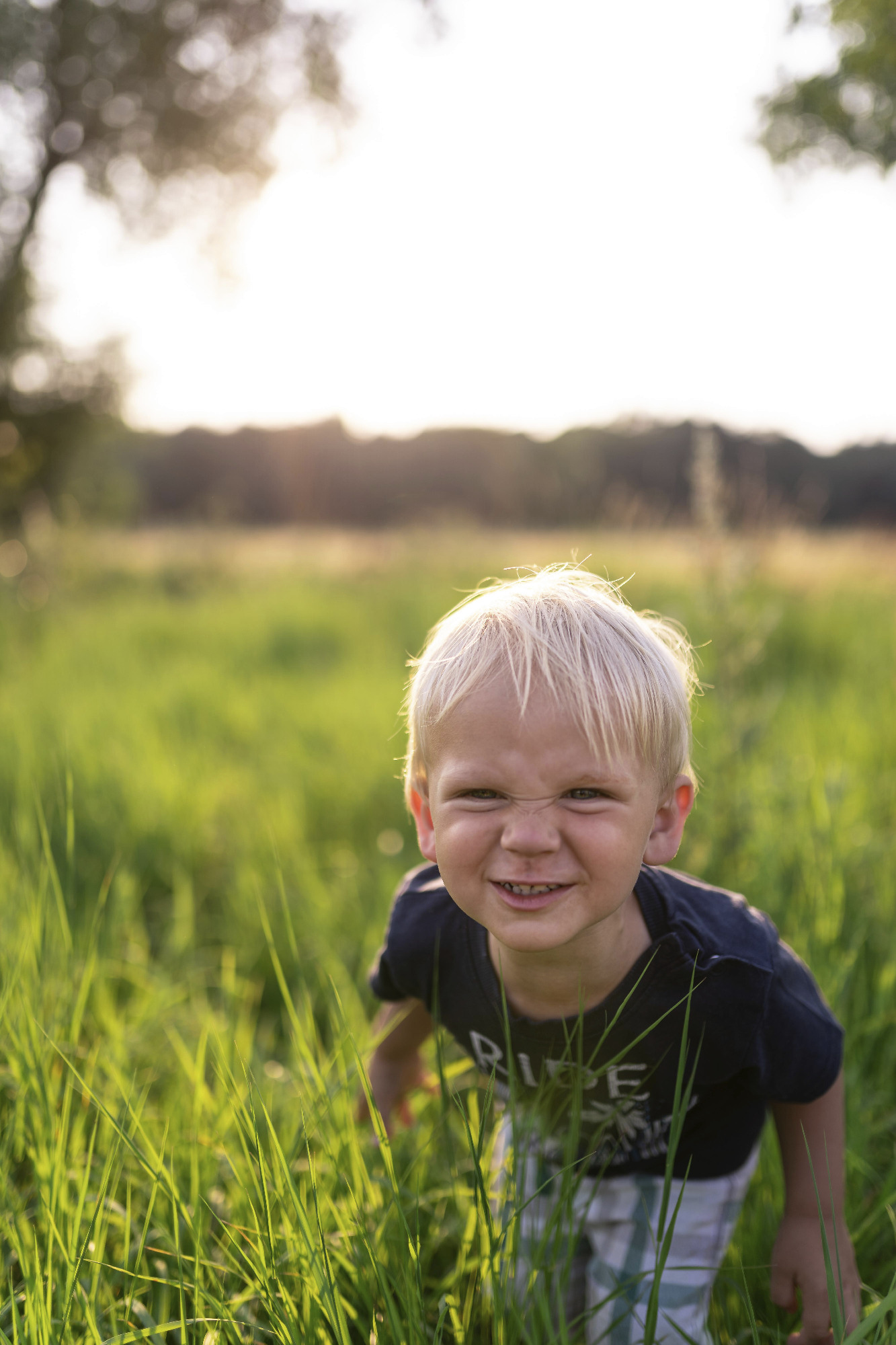 Uśmiechnięty chłopiec z blond włosami stoi w wysokiej, zielonej trawie na tle zachodzącego słońca.