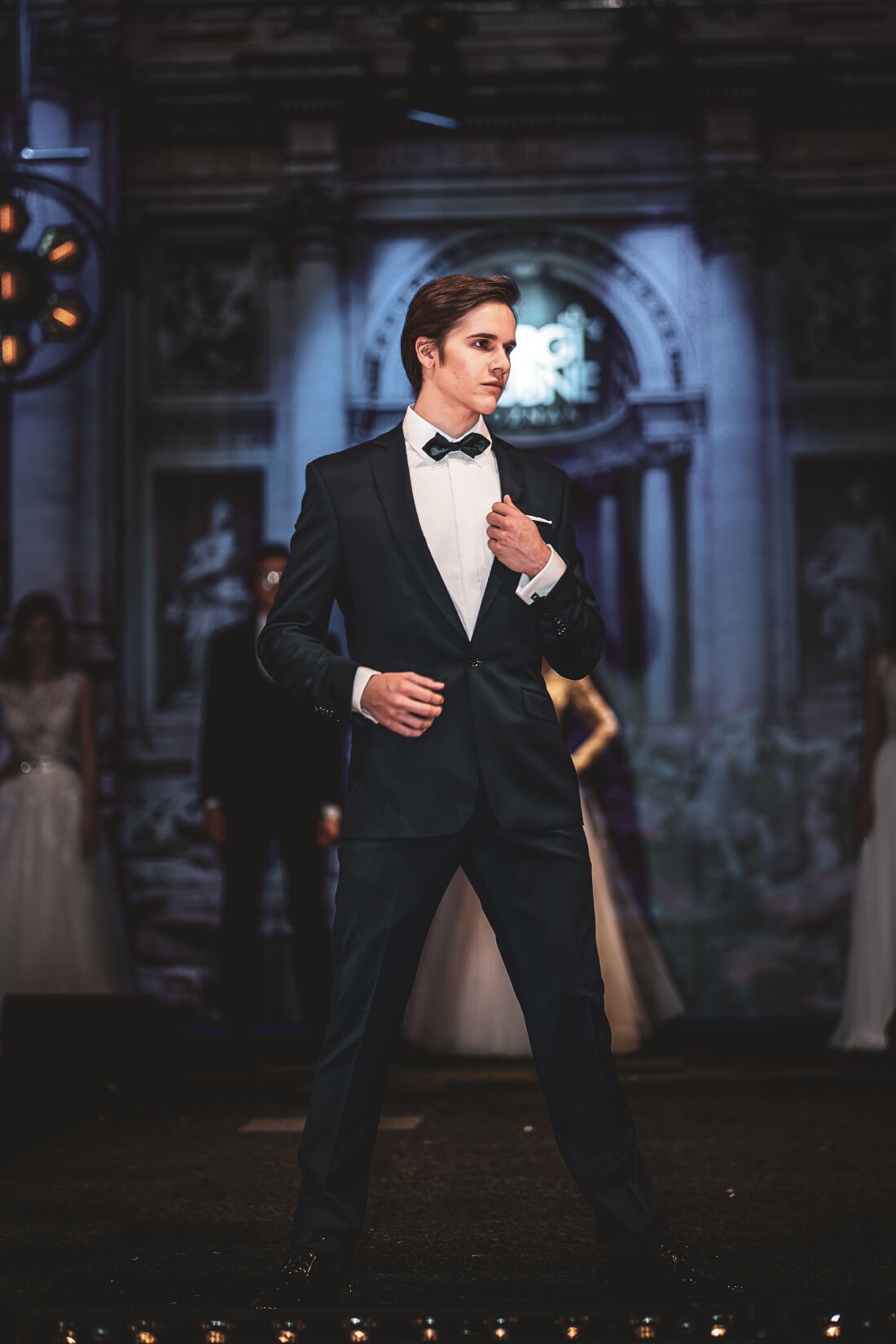 Model w granatowym smokingu i muszce na wybiegu podczas pokazu mody ślubnej, oświetlenie sceniczne i dekoracje w tle.