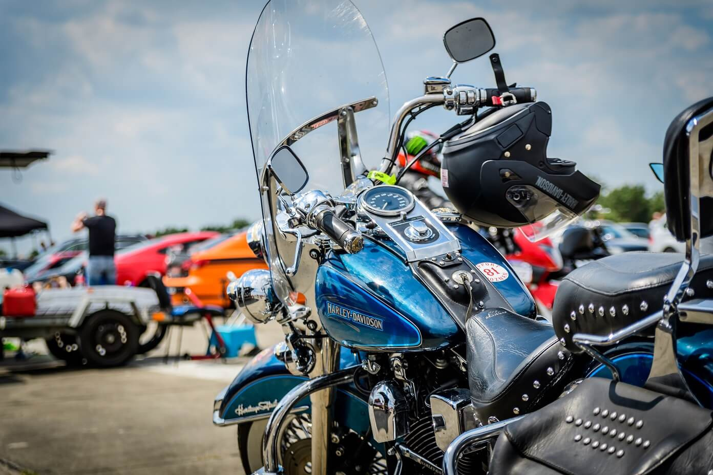 Błyszczący, niebieski motocykl Harley-Davidson z szybą i chromowanymi elementami, kask obok, na tle parkingu z samochodami i osobą robiącą zdjęcie.