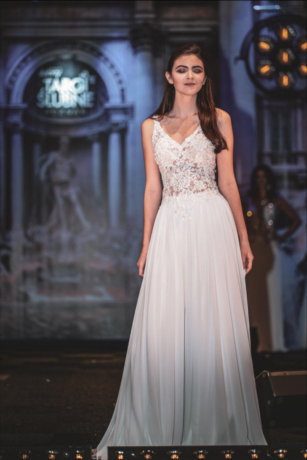 Modelka prezentuje białą suknię ślubną z koronkową górą na tle sceny z napisem 'Happy Targi Slubne'.