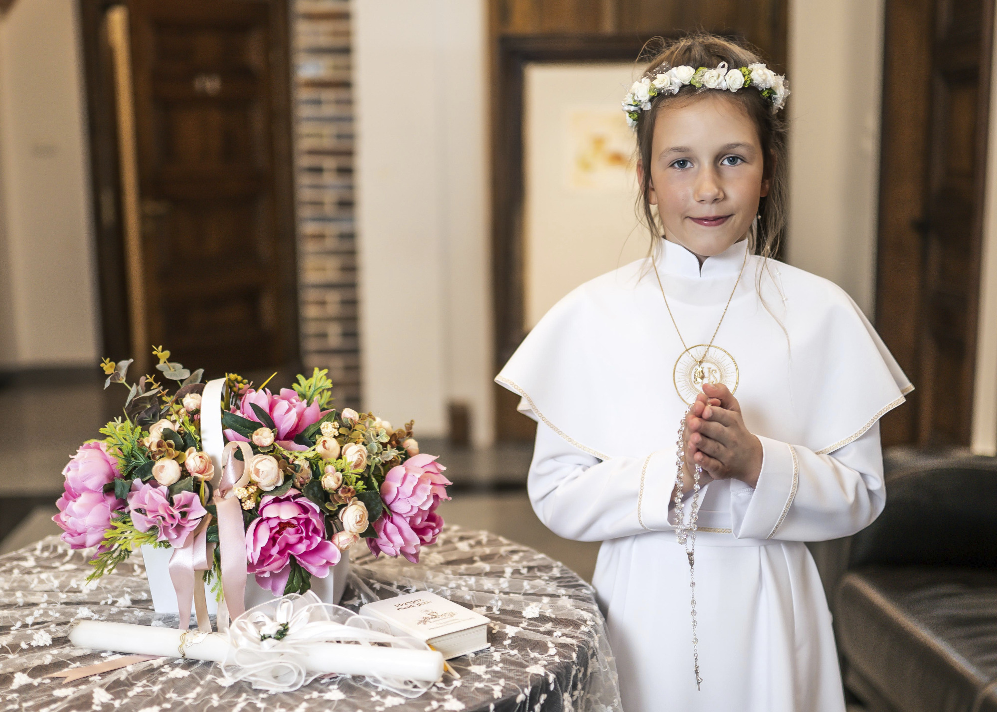 Dziewczynka w białej albie i wianku, trzymająca różaniec, obok bukiet kwiatów i świeca na stole z koronkowym obrusem, w tle drzwi.