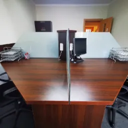 Dwa stanowiska pracy z ciemnobrązowymi biurkami oddzielone szklaną przegrodą, wyposażone w monitory, metalowe organizery na dokumenty i białe telefony na ścianie.