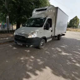 Biały samochód dostawczy Iveco z zabudową kontenerową zaparkowany na brukowanej drodze, w tle drzewa i plac zabaw.