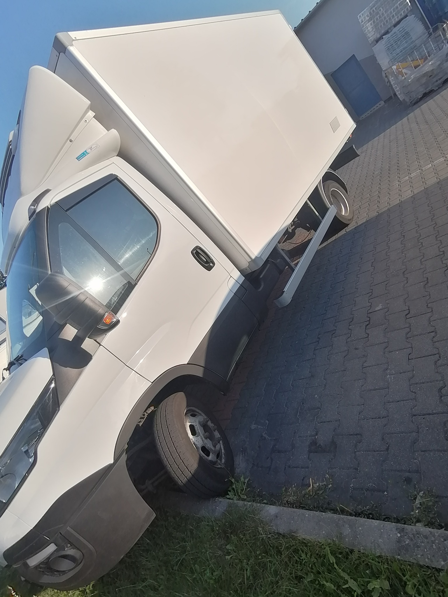 Biały samochód dostawczy z zabudową kontenerową, zaparkowany na kostce brukowej, widok z przodu i boku, z widocznym stopniem wejściowym.