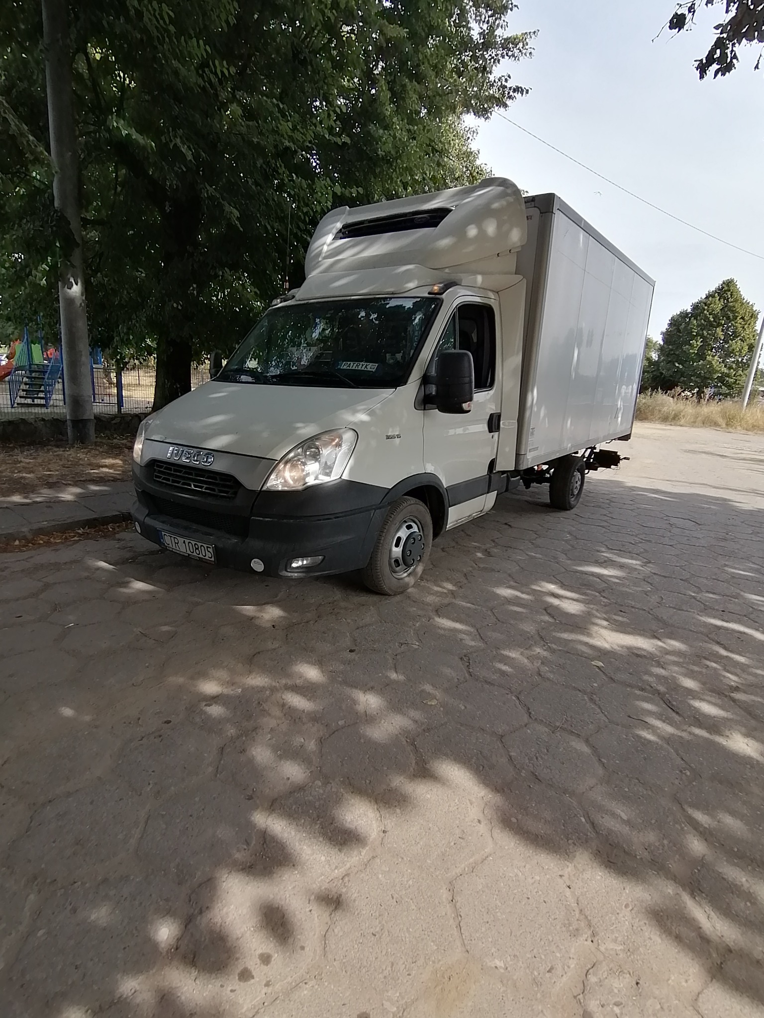 Biały samochód dostawczy Iveco z zabudową kontenerową zaparkowany na brukowanej drodze, w tle drzewa i plac zabaw.