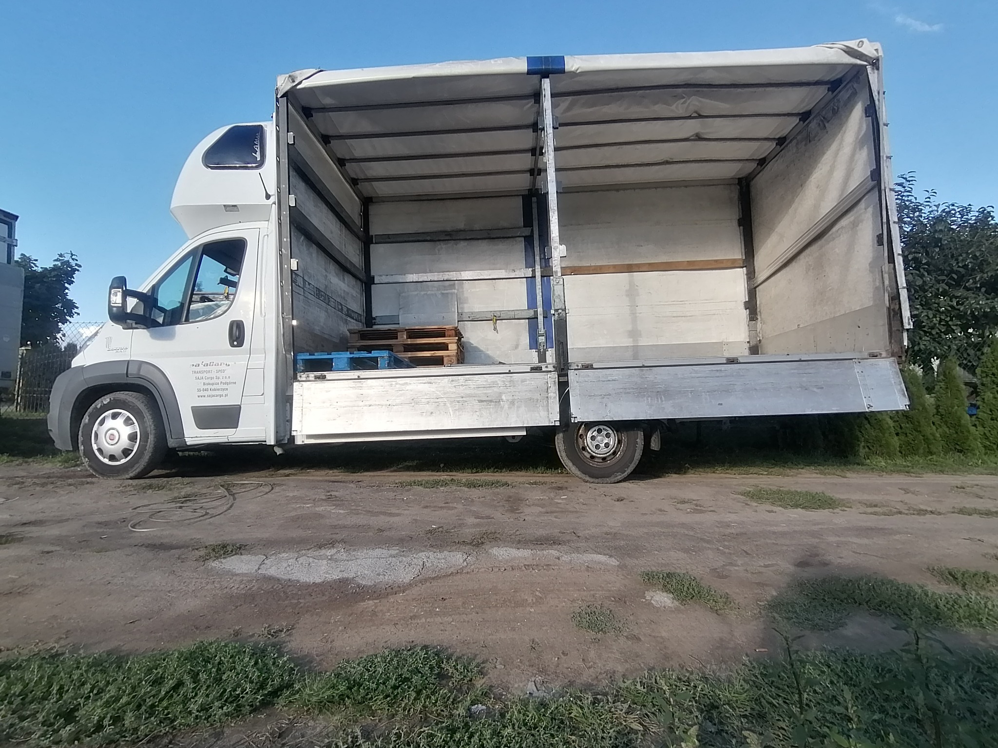 Otwarty biały bus transportowy z paletami wewnątrz, widok z boku na nierównym terenie.