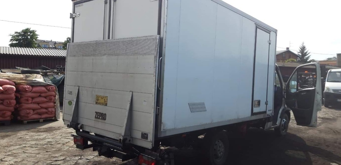 Tył białego samochodu dostawczego z otwartymi drzwiami i windą załadowczą, obok sterta worków na palecie.