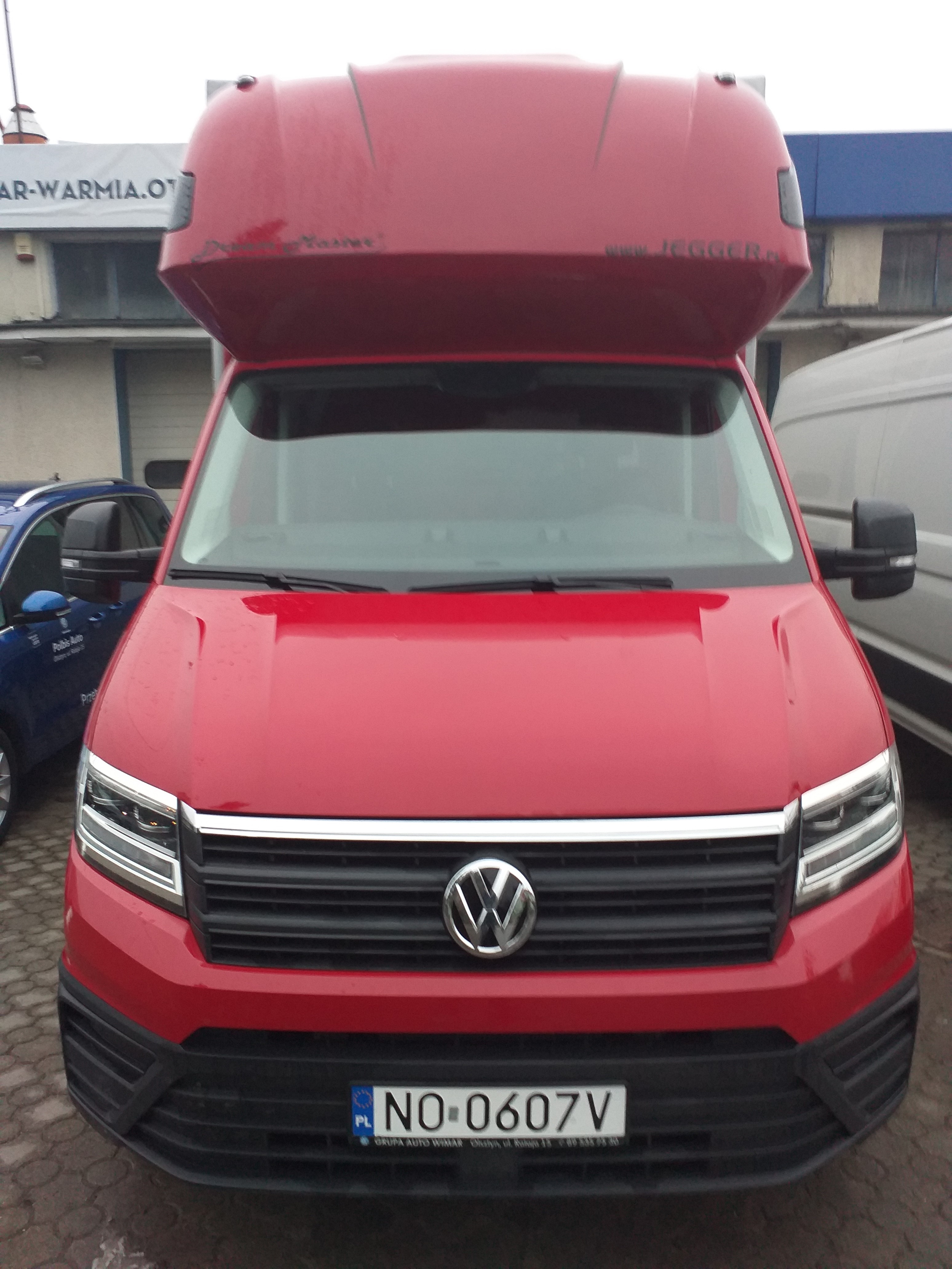 Czerwony bus Volkswagen Crafter z wysokim dachem, widok z przodu, tablica rejestracyjna NO-0607V, na dachu widoczny napis 'www.JEGGER.pl'.