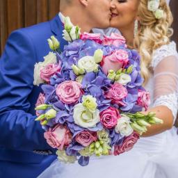 Beata Borgus BB Florist Pracownia Florystyczna Beata Borgus - Para młoda całuje się za bukietem ślubnym z różowych róż, białych eustom i niebieskich hortensji na tle ceglanej ściany.