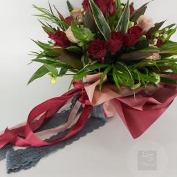 Beata Borgus BB Florist Pracownia Florystyczna Beata Borgus - Bukiet z czerwonych i różowych róż, lilii i zielonych liści, owinięty w papier w odcieniach różu i czerwieni, ozdobiony wstążkami w podobnych kolorach i szarą koronką.