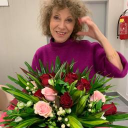 Beata Borgus BB Florist Pracownia Florystyczna Beata Borgus - Uśmiechnięta kobieta z kręconymi włosami w fuksjowym swetrze prezentuje bukiet różowych i czerwonych róż z zielonymi liśćmi na tle białej ściany z gaśnicą.