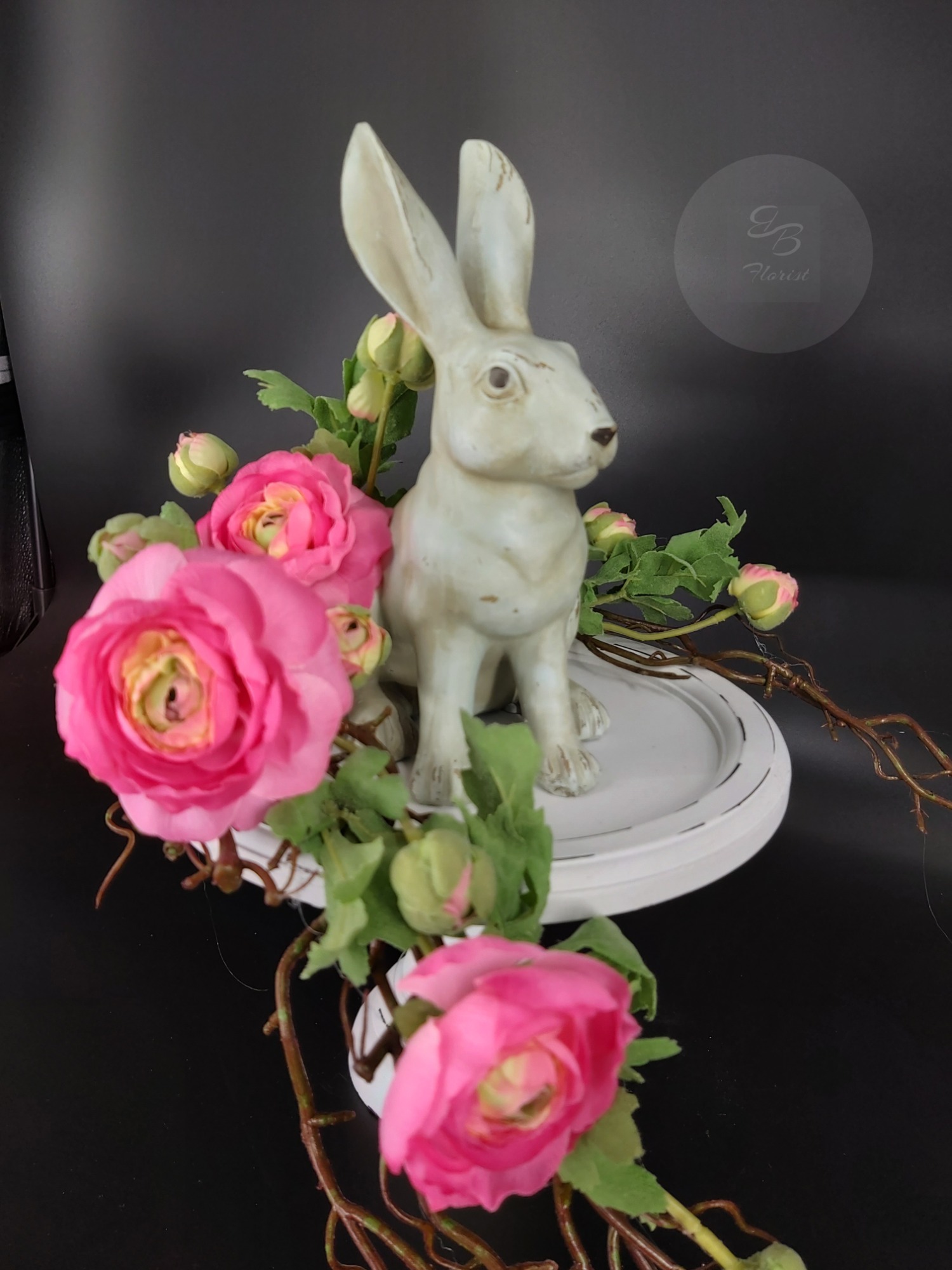 Kompozycja florystyczna z ceramicznym zającem, różowymi kwiatami i gałązkami na białej podstawie, na czarnym tle.