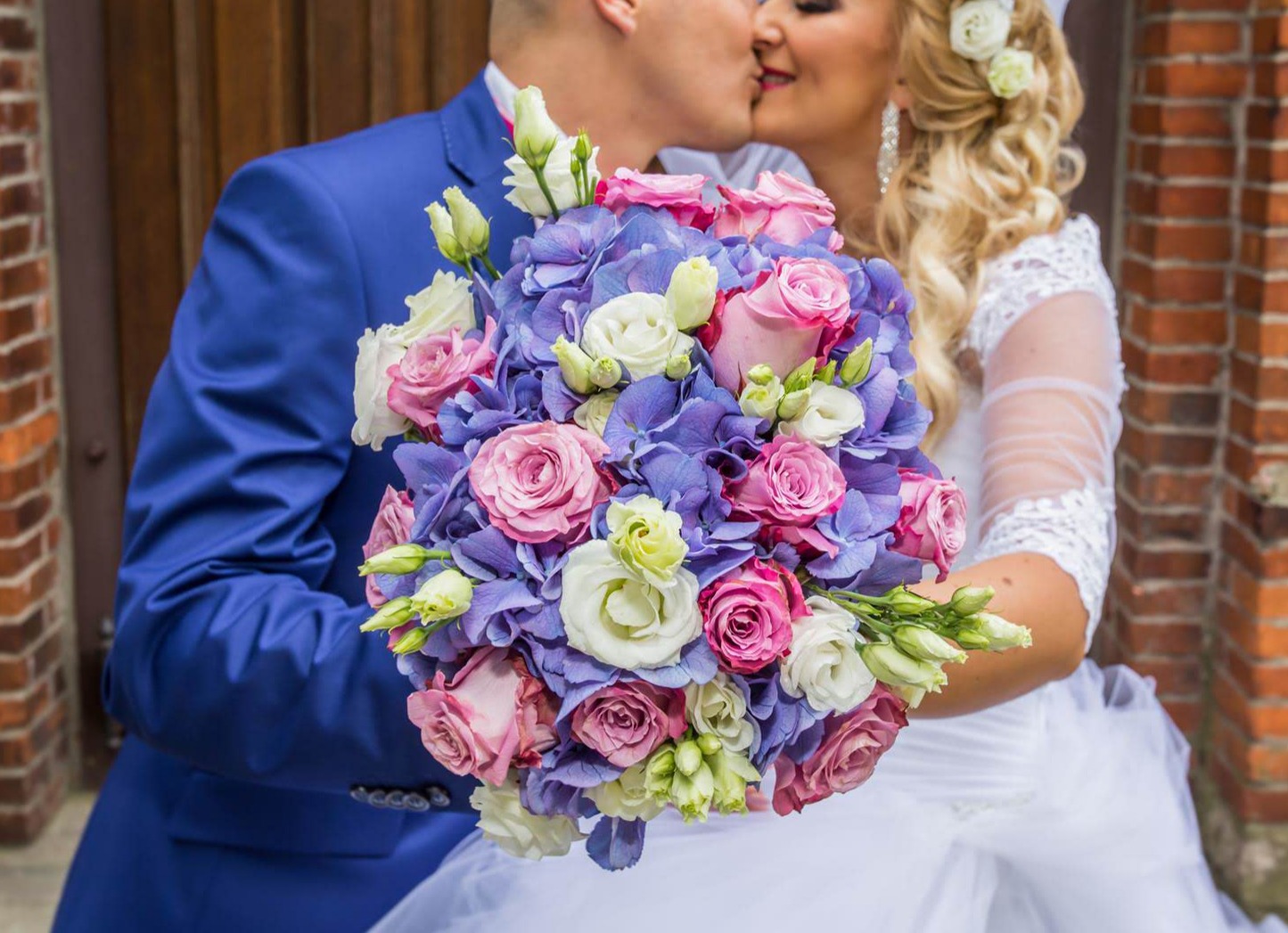 Para młoda całuje się za bukietem ślubnym z różowych róż, białych eustom i niebieskich hortensji na tle ceglanej ściany.