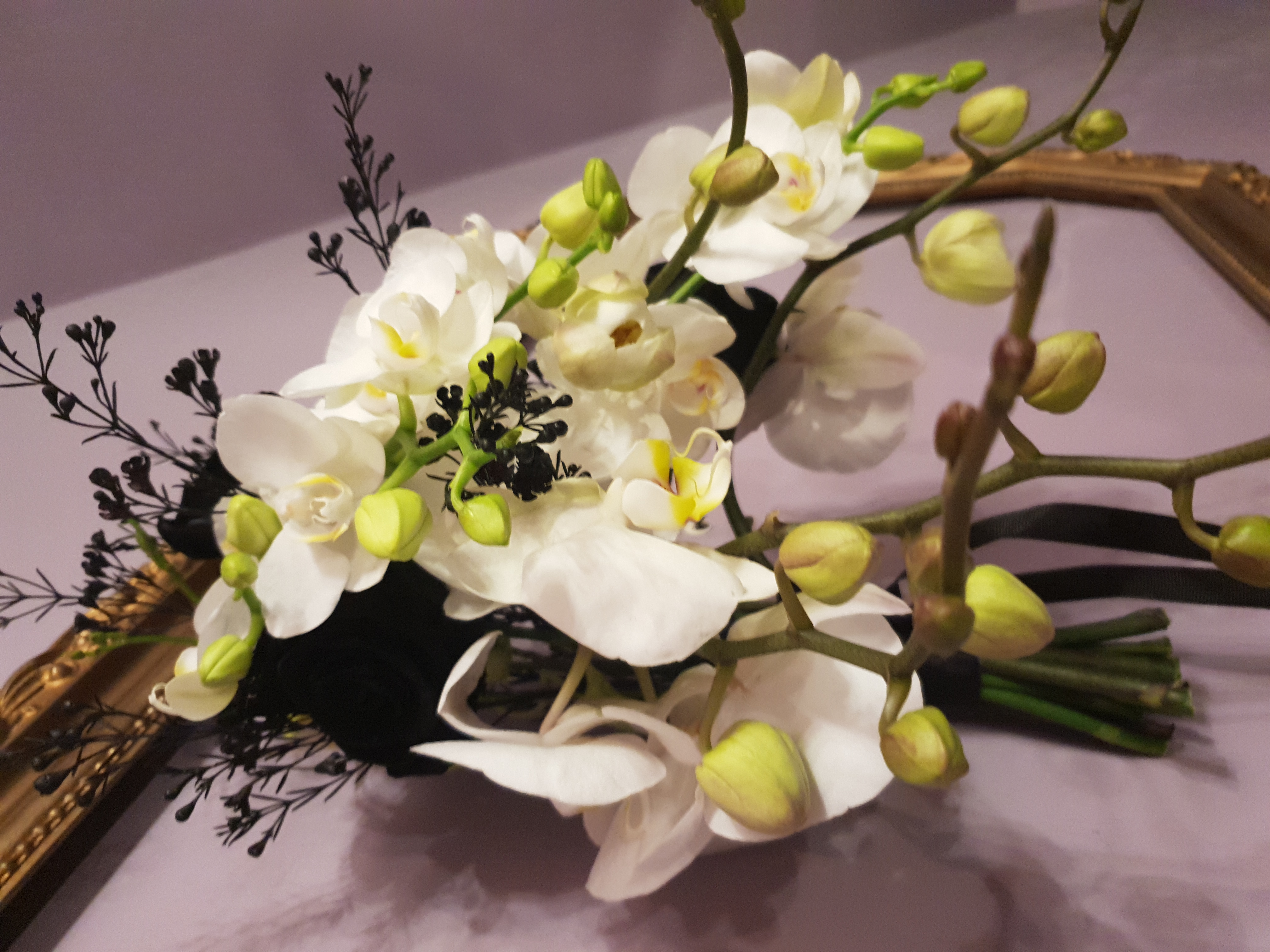 Elegancki bukiet ślubny z białych orchidei i czarnych gałązek, owinięty czarną wstążką, na tle złotego, ozdobnego obramowania.