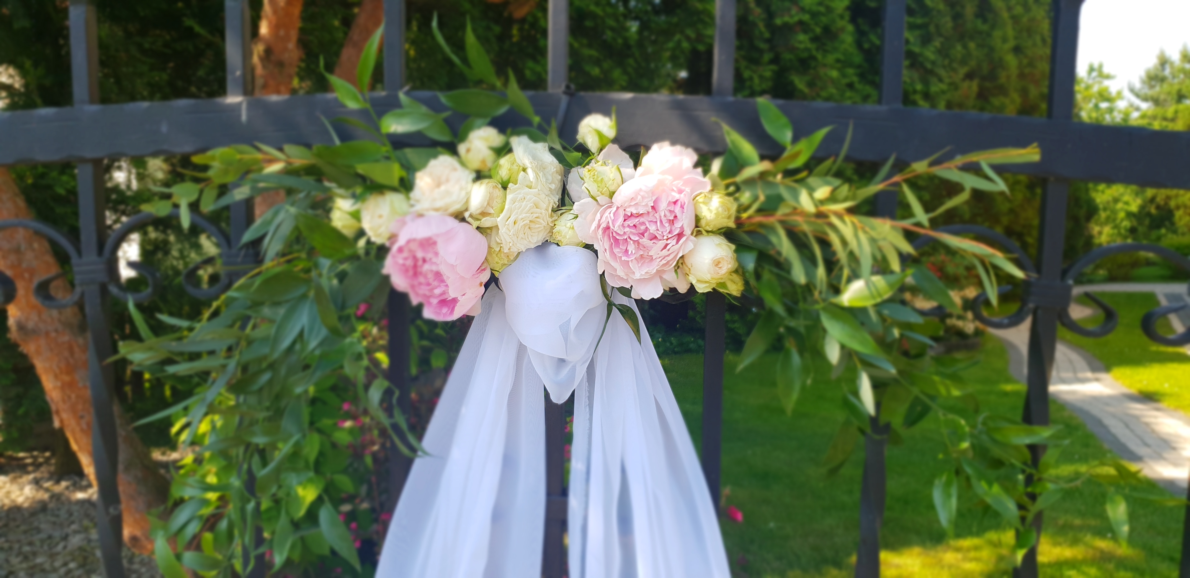 Dekoracja ślubna z białych i różowych kwiatów oraz zwiewnej tkaniny, zamocowana na metalowym ogrodzeniu z widokiem na zielony trawnik i drzewa w tle.