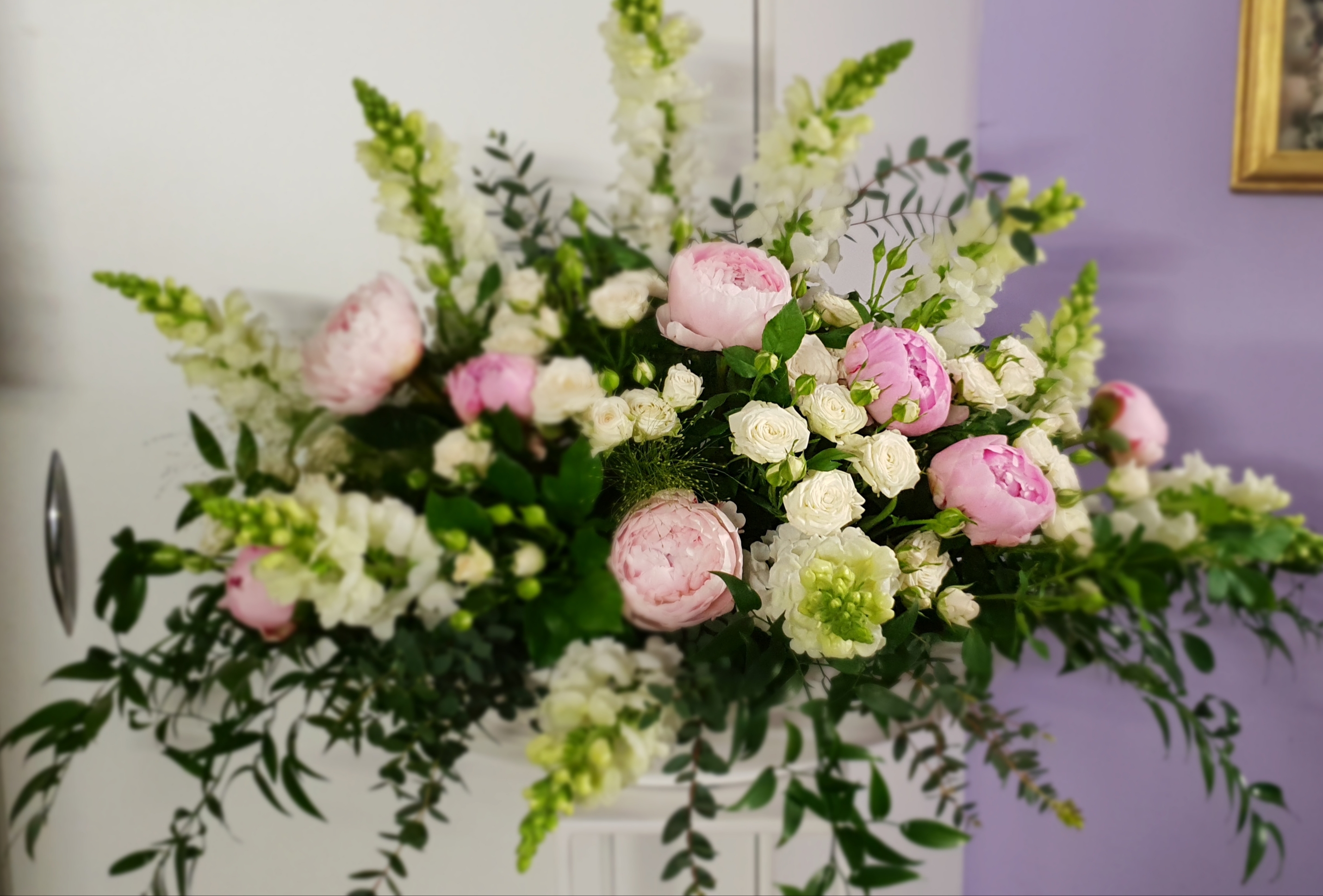 Kompozycja kwiatowa z różowych piwonii, białych róż i zielonych dodatków, umieszczona na białym tle, z fioletową ścianą i złotą ramką obrazu w tle.