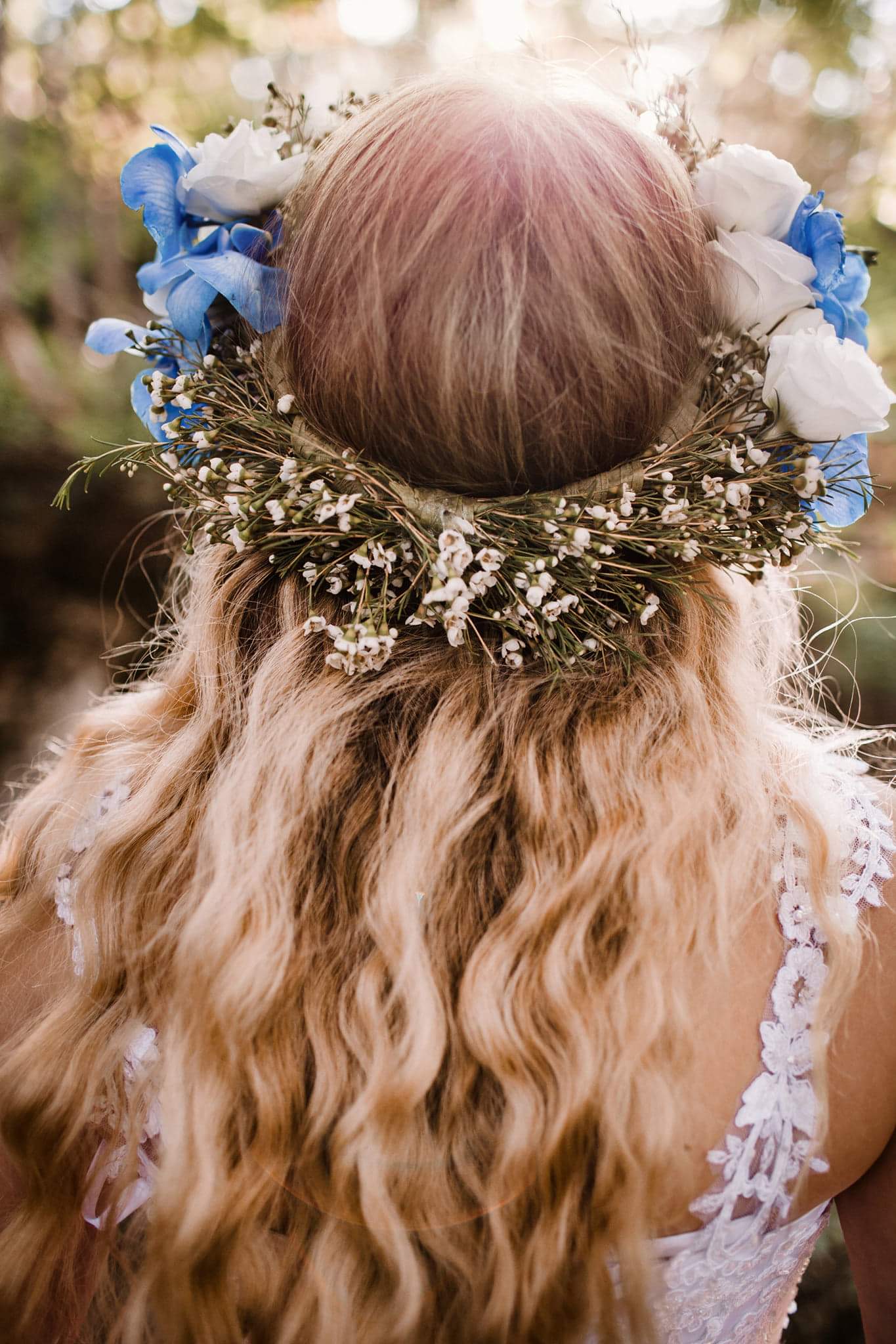 Tył głowy kobiety z blond, falowanymi włosami, ozdobionej wiankiem z niebieskich i białych kwiatów oraz drobnymi białymi kwiatuszkami, widzianej z bliska, z lekkim prześwietleniem słonecznym.