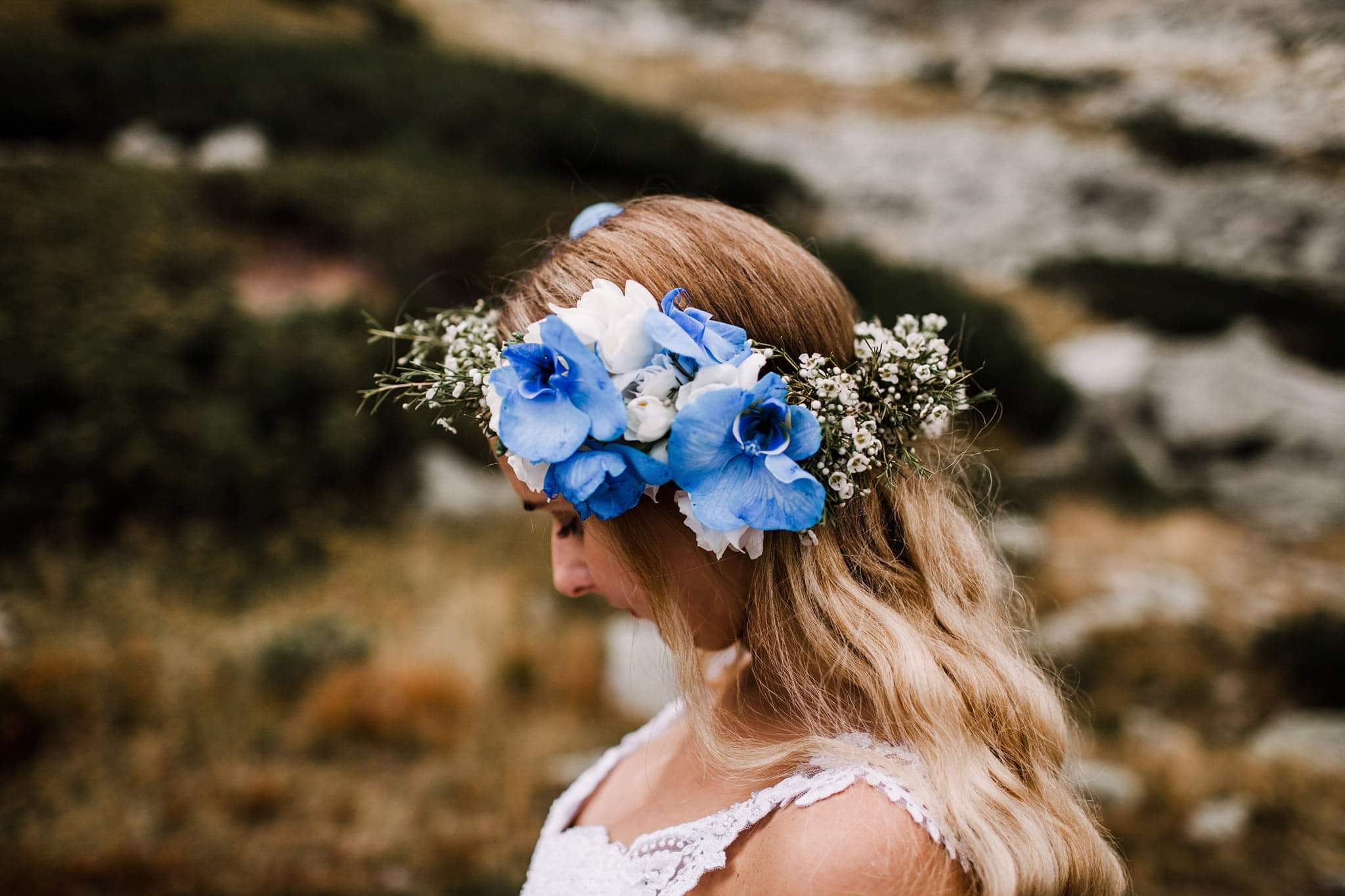 Panna młoda z blond włosami i wiankiem z niebieskich i białych kwiatów na głowie, z profilu, z delikatną koronkową sukienką.