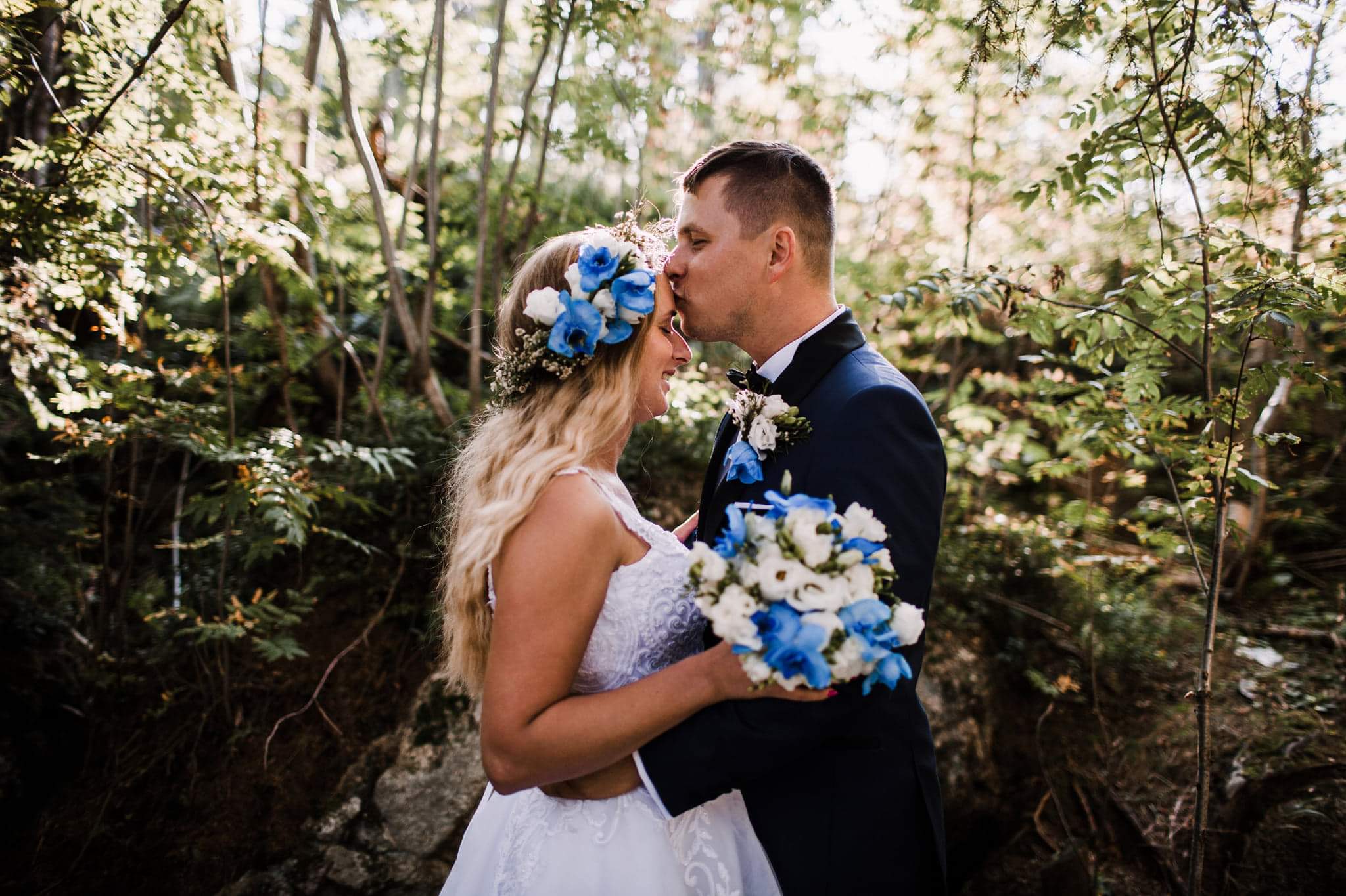 Panna młoda w białej sukni i wianku z niebieskich kwiatów, obejmowana przez pana młodego w granatowym garniturze, który całuje ją w czoło na tle leśnej scenerii.