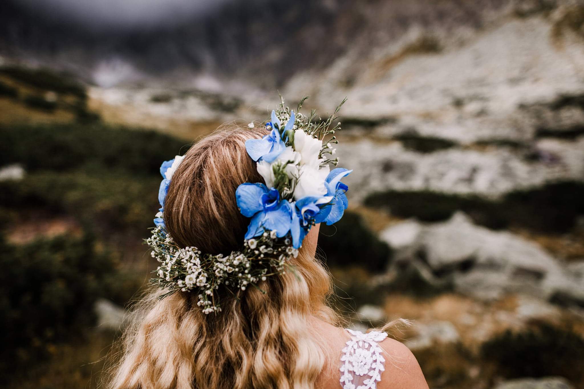 Panna młoda z blond włosami i wiankiem z białych i niebieskich kwiatów na tle górskiego krajobrazu.