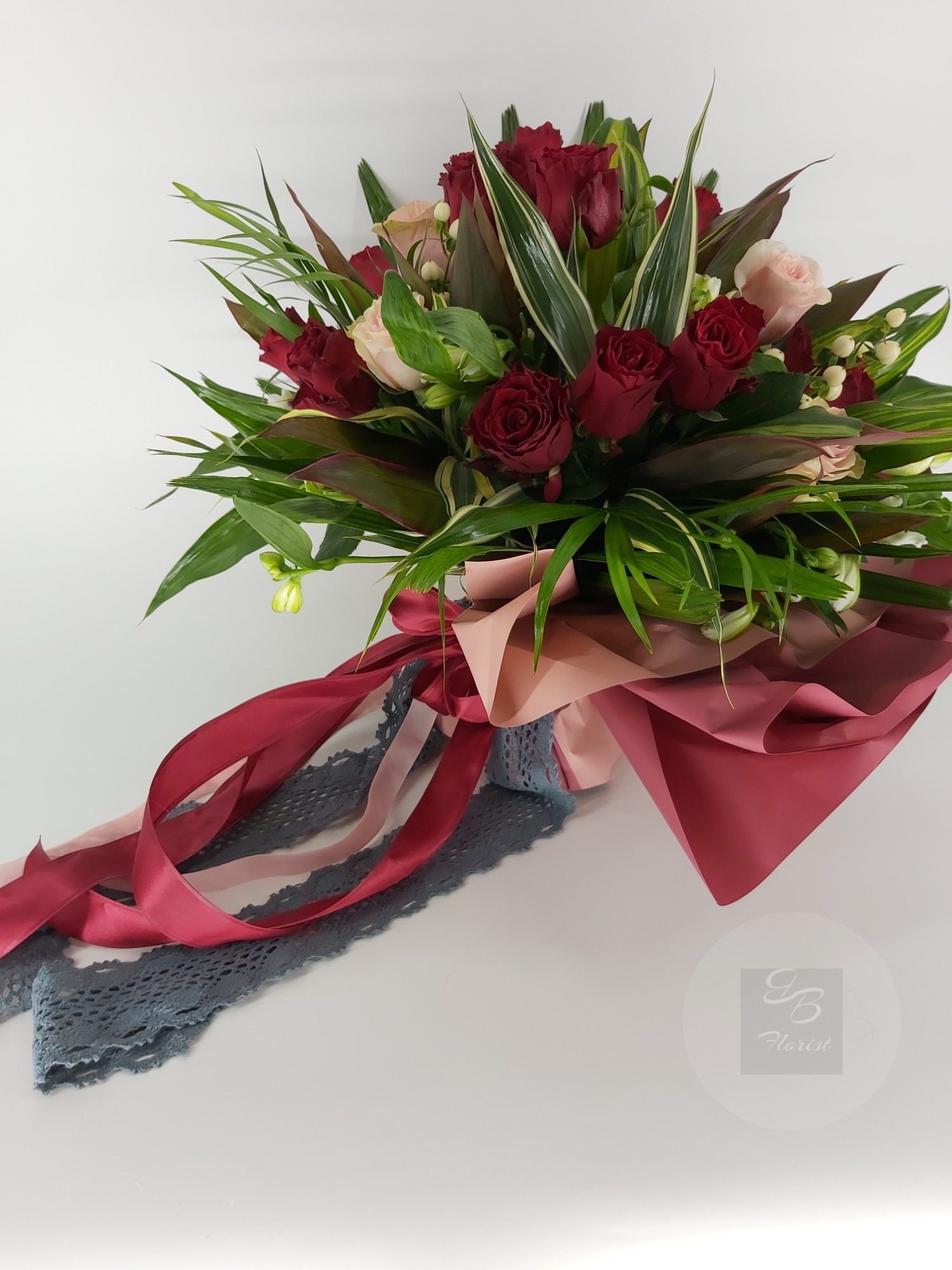 Bukiet z czerwonych i różowych róż, lilii i zielonych liści, owinięty w papier w odcieniach różu i czerwieni, ozdobiony wstążkami w podobnych kolorach i szarą koronką.