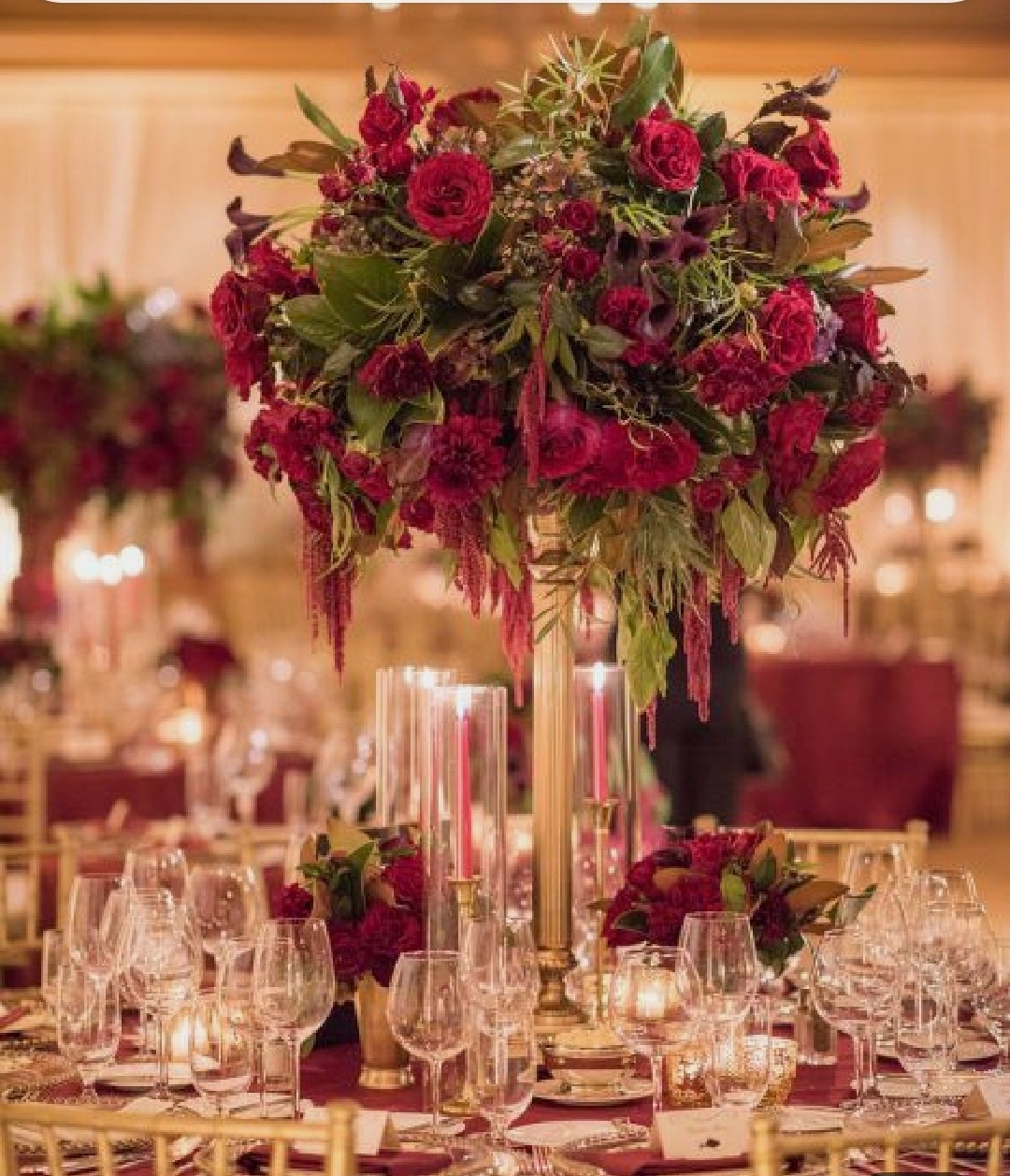 Uroczysta dekoracja stołu z wysokim, bogatym bukietem z czerwonych róż i amarantusa w złotym wazonie, otoczona szklanymi świecznikami i kryształowymi kieliszkami na tle sali bankietowej.