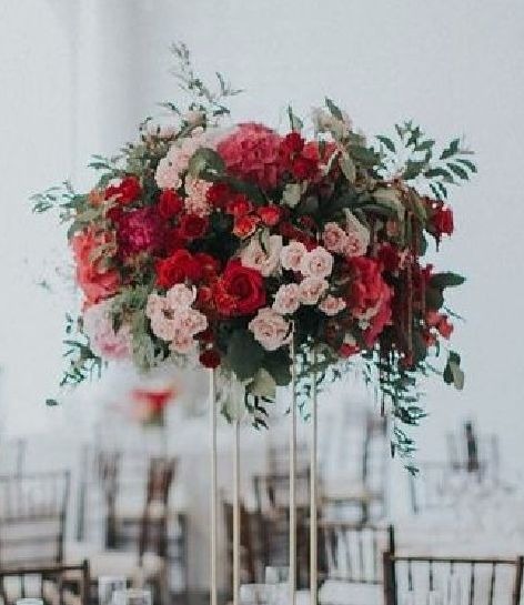 Kompozycja kwiatowa z czerwonych i różowych róż, piwonii i zielonych liści na wysokim, złotym stojaku, ustawiona na stole z nakryciami w tle sali bankietowej.