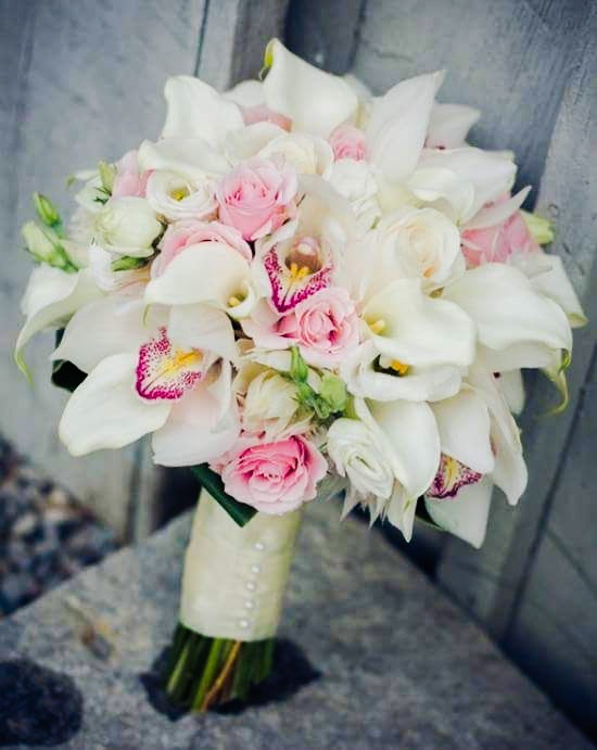 Bukiet ślubny z białych kalii i różowych róż, owinięty kremową wstążką z perłowymi akcentami, na tle szarej, teksturowanej powierzchni.
