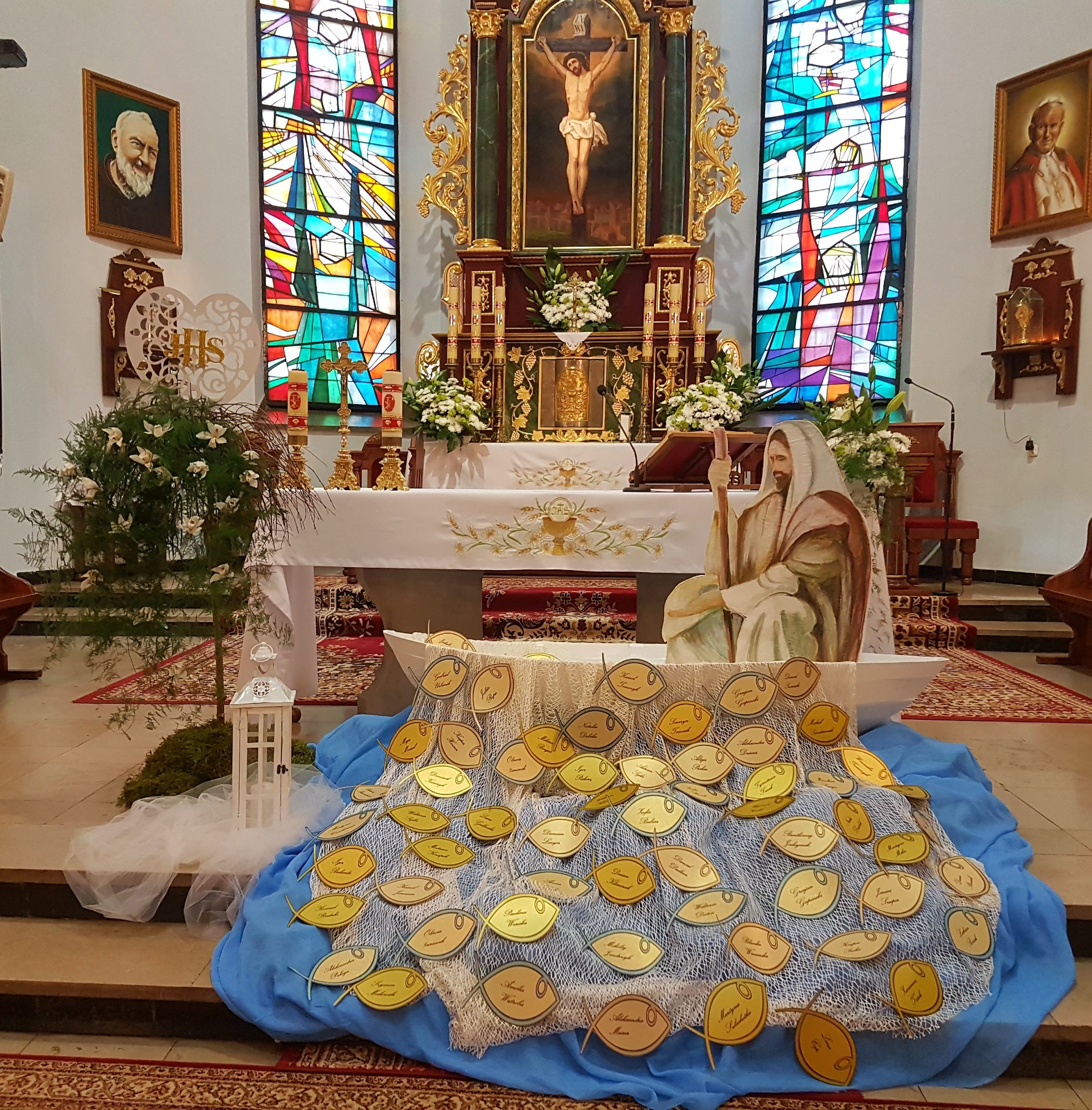 Dekoracja w kościele z okazji uroczystości, centralnie umieszczona drewniana łódka wypełniona złotymi rybkami z imionami, w tle ołtarz i witraże.