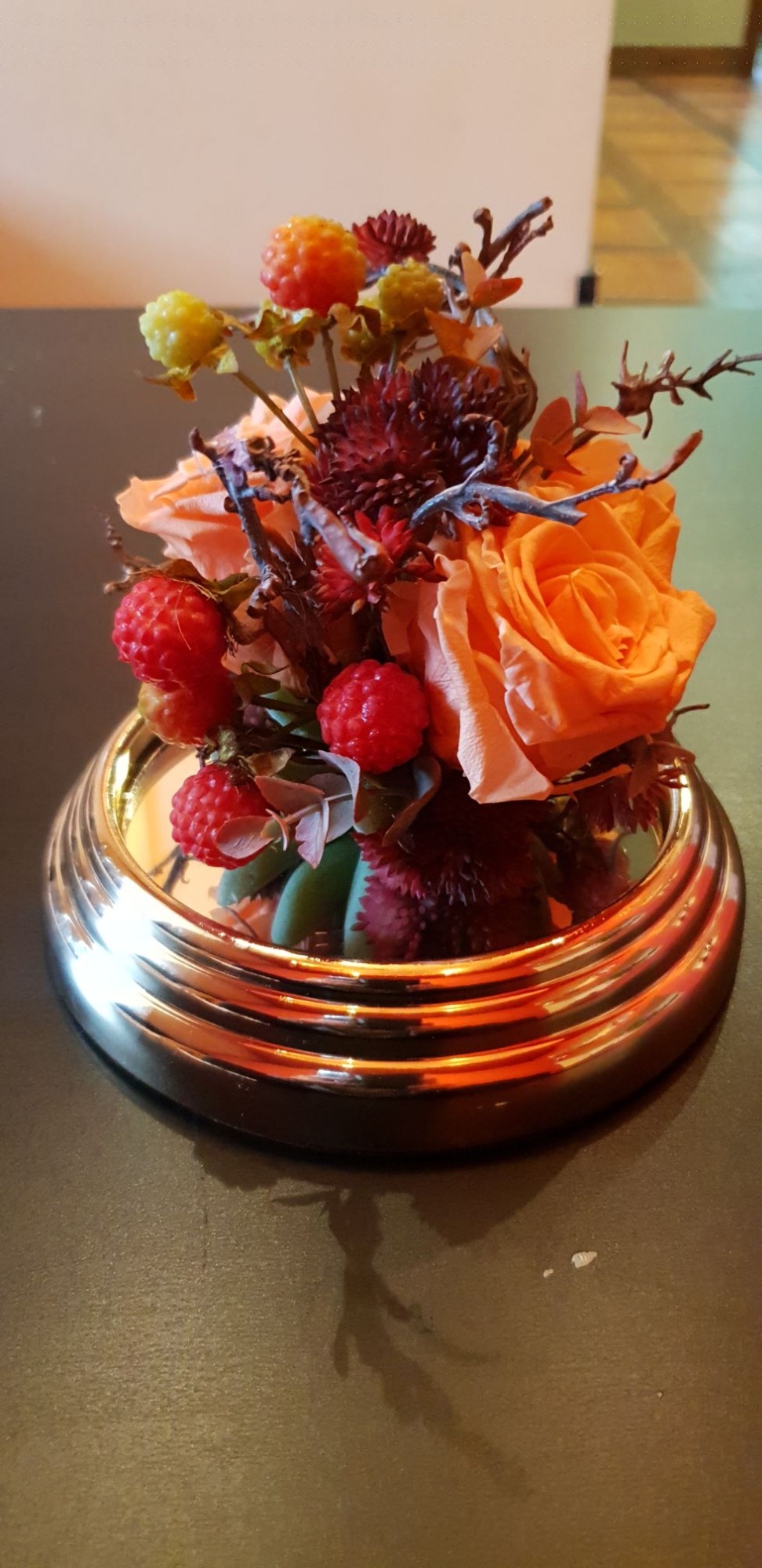 Kompozycja kwiatowa z pomarańczowych róż, czerwonych koralików i brązowych gałązek w okrągłej, złotej ramce na ciemnym tle.