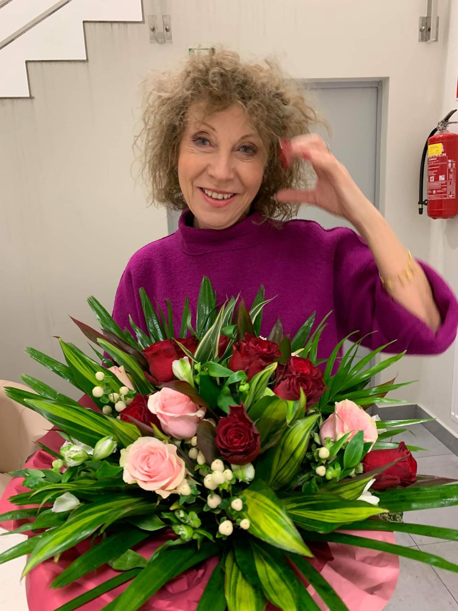 Uśmiechnięta kobieta z kręconymi włosami w fuksjowym swetrze prezentuje bukiet różowych i czerwonych róż z zielonymi liśćmi na tle białej ściany z gaśnicą.