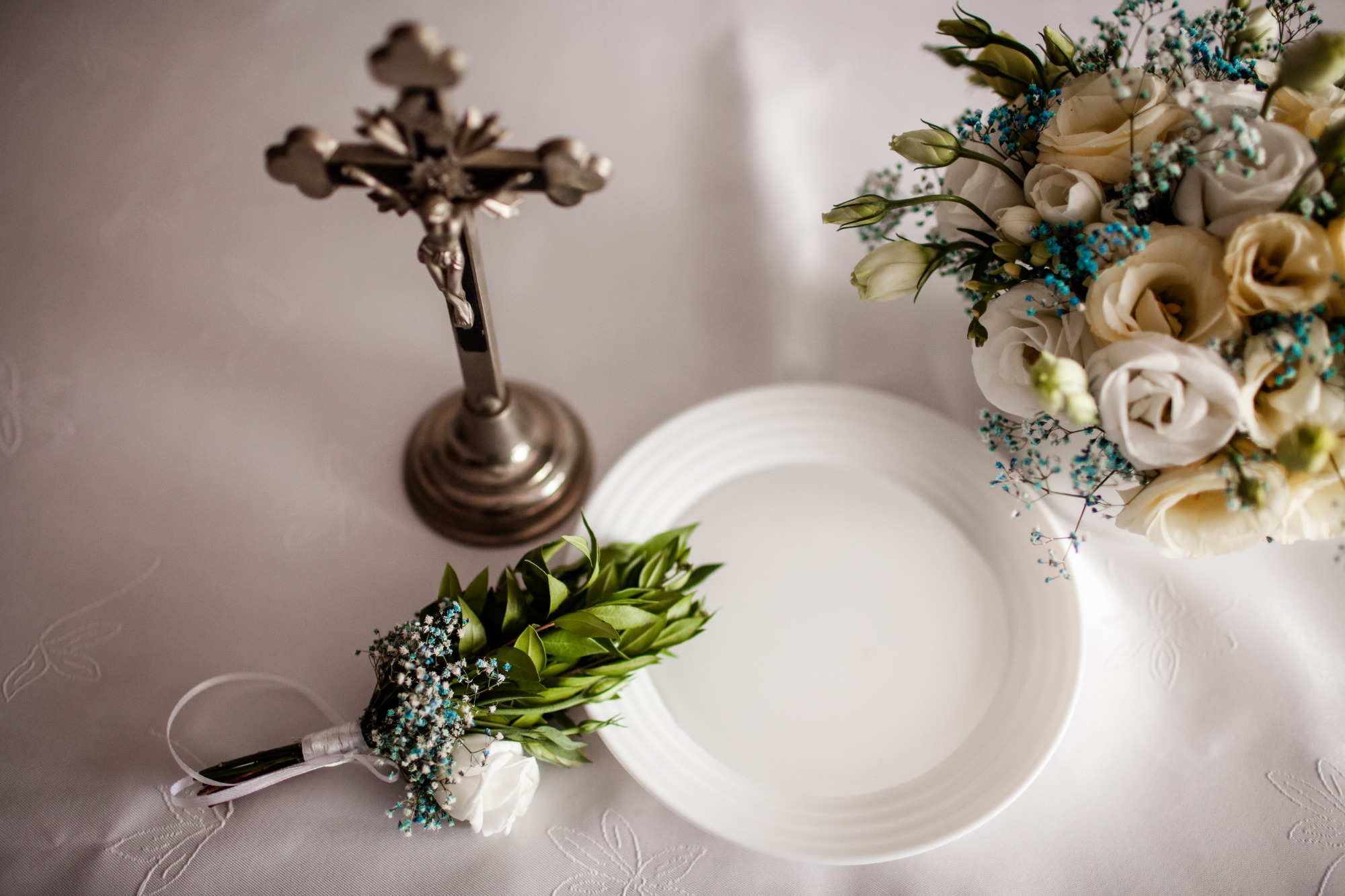 Przygotowania do ceremonii ślubnej: srebrny krucyfiks, bukiet ślubny z białych róż i niebieskich akcentów, butonierka z zielonych liści i białą różą, pusty biały talerz na białym obrusie...
