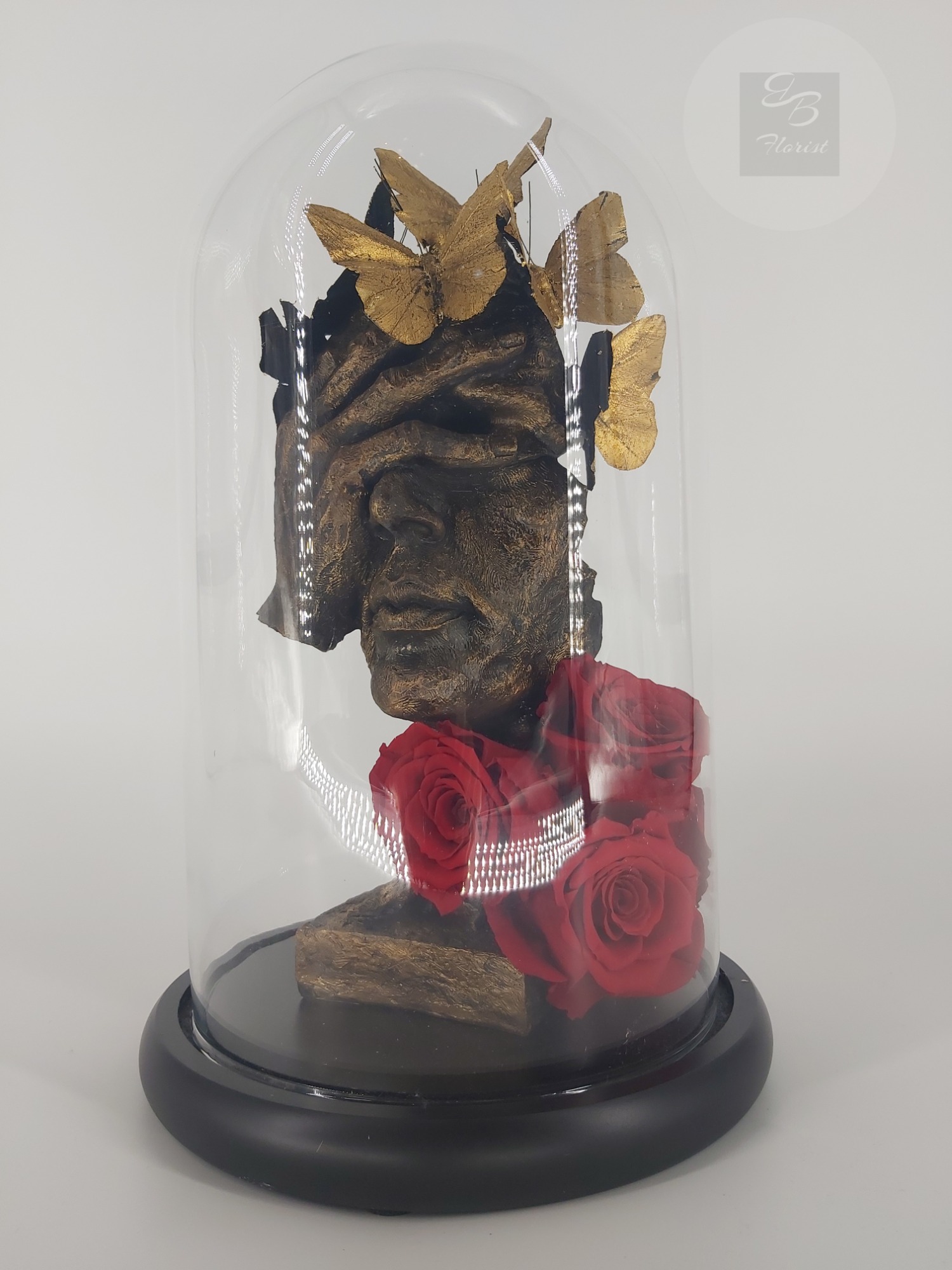 Szklana kopuła z rzeźbą głowy z zasłoniętymi oczami, złotymi motylami i czerwonymi różami.