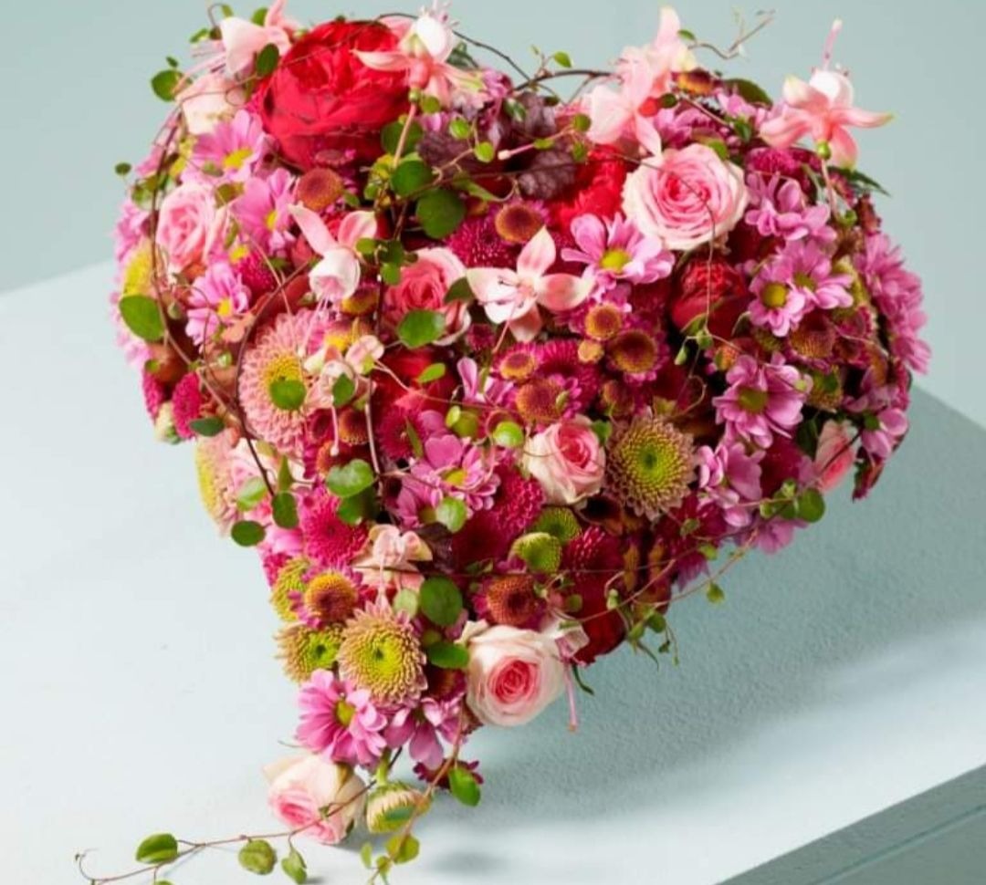 Kompozycja kwiatowa w kształcie serca z róż, chryzantem i drobnych kwiatów w odcieniach różu i czerwieni, ułożona na jasnoniebieskim tle.