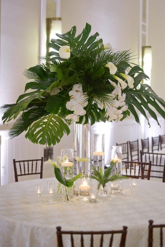 Dekoracja stołu z wysoką kompozycją z liści monstery, białych kalii i storczyków w szklanych wazonach z pływającymi świecami, ustawiona na białym obrusie w sali bankietowej.