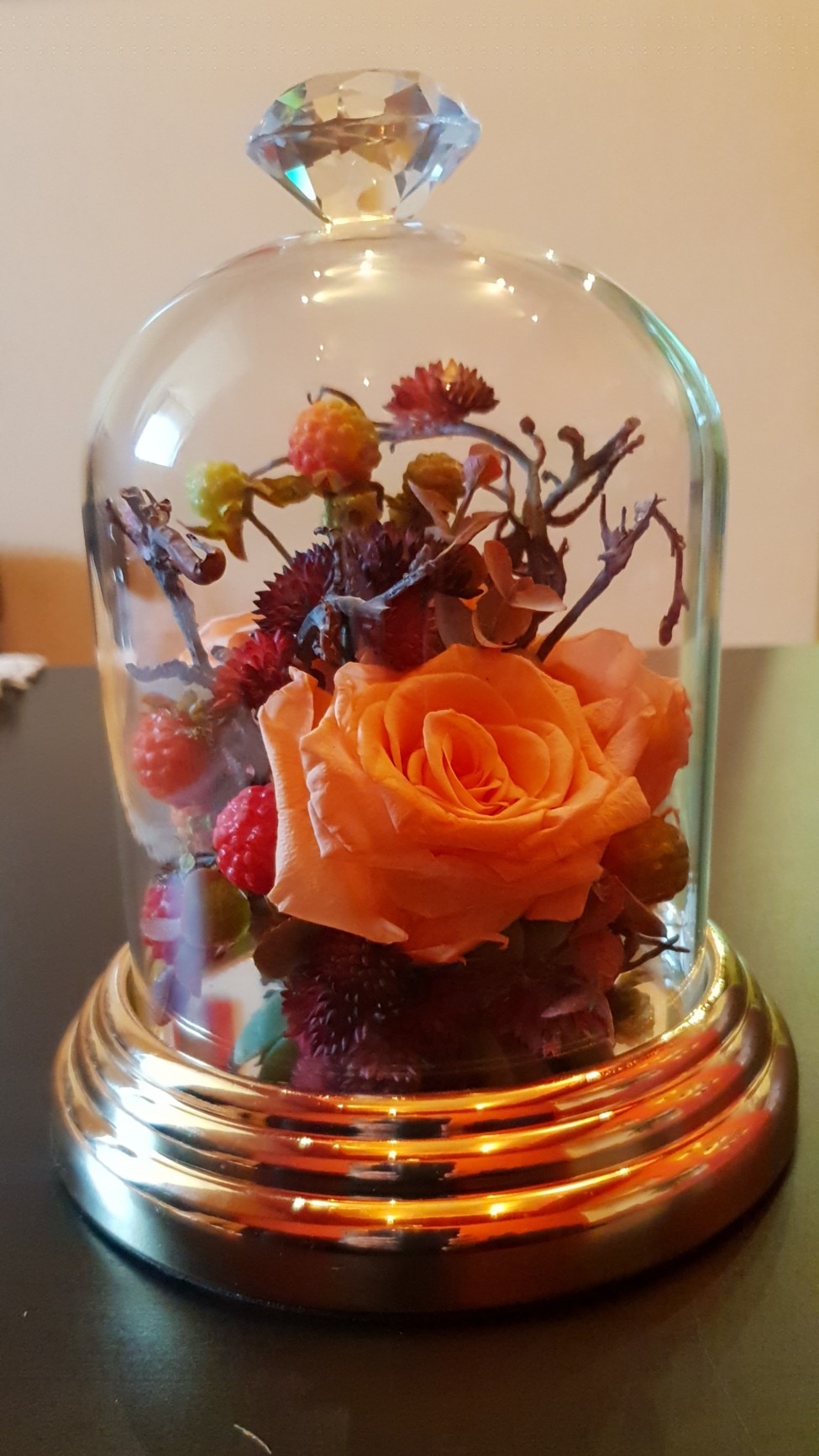 Dekoracja stołu: pomarańczowa róża i suszone kwiaty pod szklanym kloszem z kryształową gałką, na złotej podstawie.