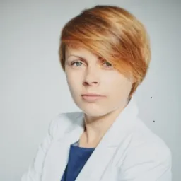 Portret kobiety z krótkimi rudymi włosami, ubrana w białą marynarkę i niebieską bluzkę, na jasnoszarym tle.