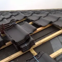 Układanie ciemnych dachówek ceramicznych na drewnianej konstrukcji dachu pokrytej membraną, widoczne stosy dachówek zabezpieczone taśmą.