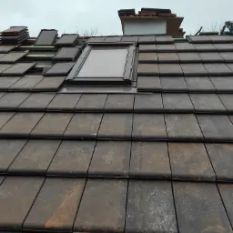 Montaż okna dachowego na skośnym dachu pokrytym ciemnymi dachówkami ceramicznymi, z widocznymi elementami konstrukcji drewnianej i ułożonymi dachówkami w tle, pochmurne niebo.