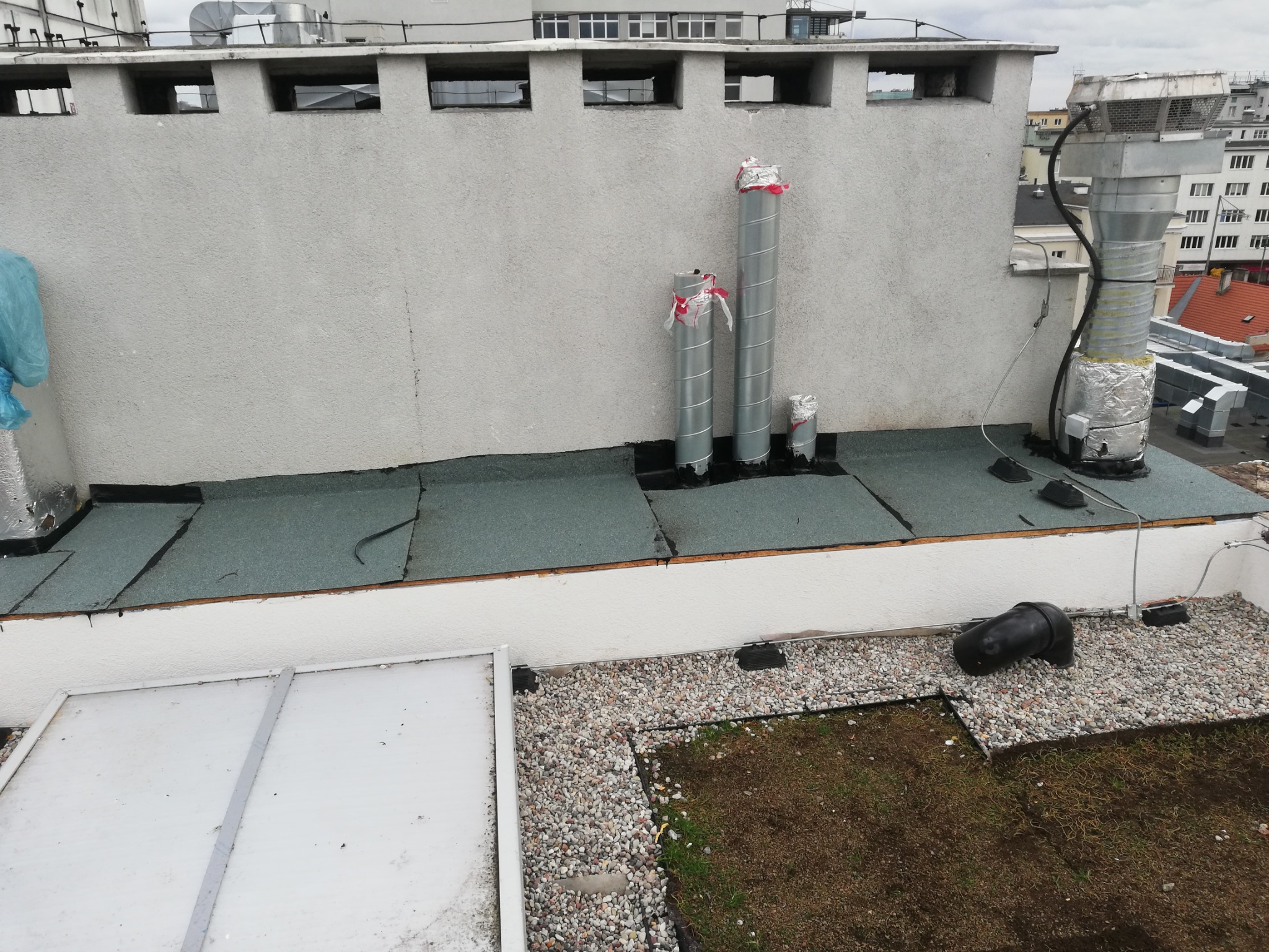 Widok z góry na płaski dach budynku z zielonym dachem, elementami wentylacyjnymi i kominami, oraz fragmentem instalacji elektrycznej.