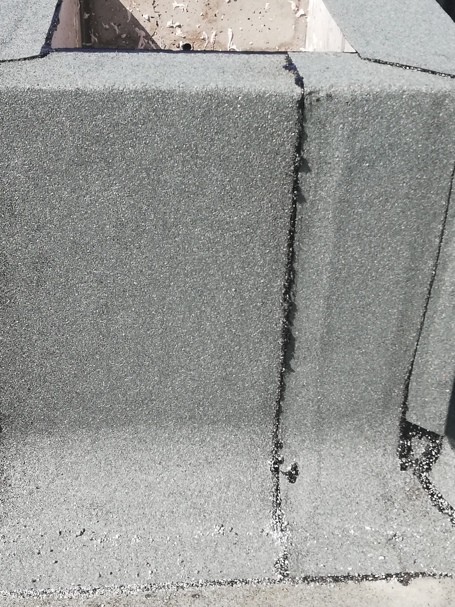 Szczegółowe ujęcie fragmentu dachu pokrytego szarą papą z widocznymi łączeniami i śladami napraw dekarskich na kominie.