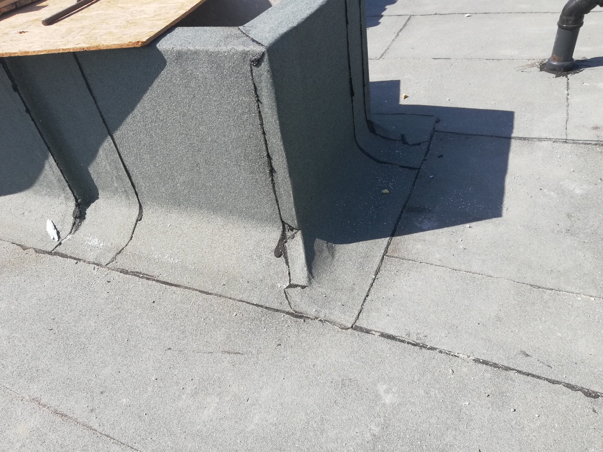 Szare, płaskie pokrycie dachowe z papy bitumicznej, obróbka komina z widocznymi łączeniami i fragmentem drewnianej płyty.