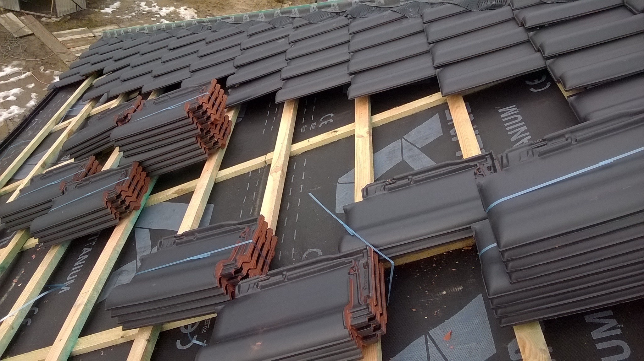 Ciemnoszare dachówki ceramiczne ułożone na łatach drewnianych, częściowo pokrywające czarną membranę dachową, przygotowane do montażu na dachu.