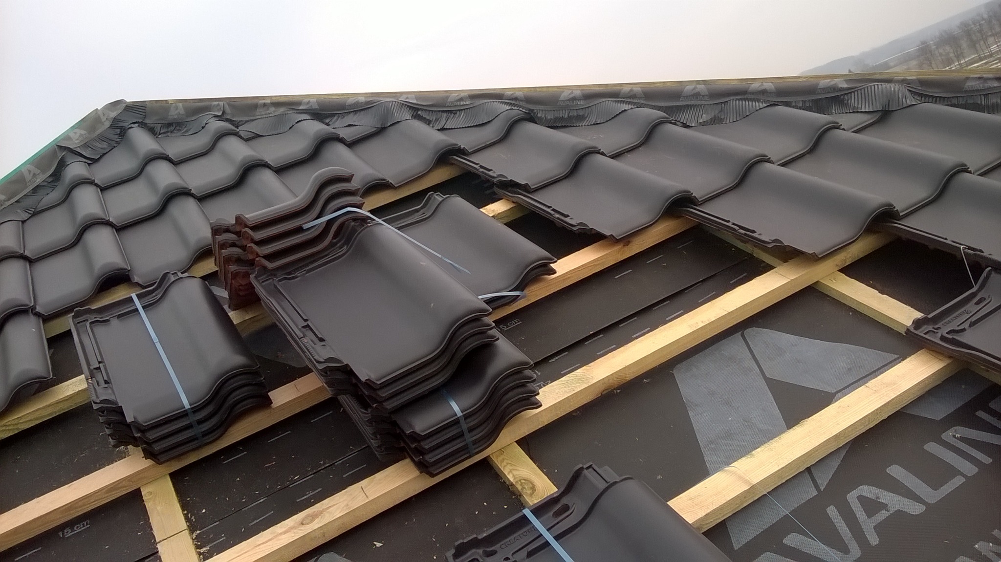 Układanie ciemnych dachówek ceramicznych na drewnianej konstrukcji dachu pokrytej membraną, widoczne stosy dachówek zabezpieczone taśmą.